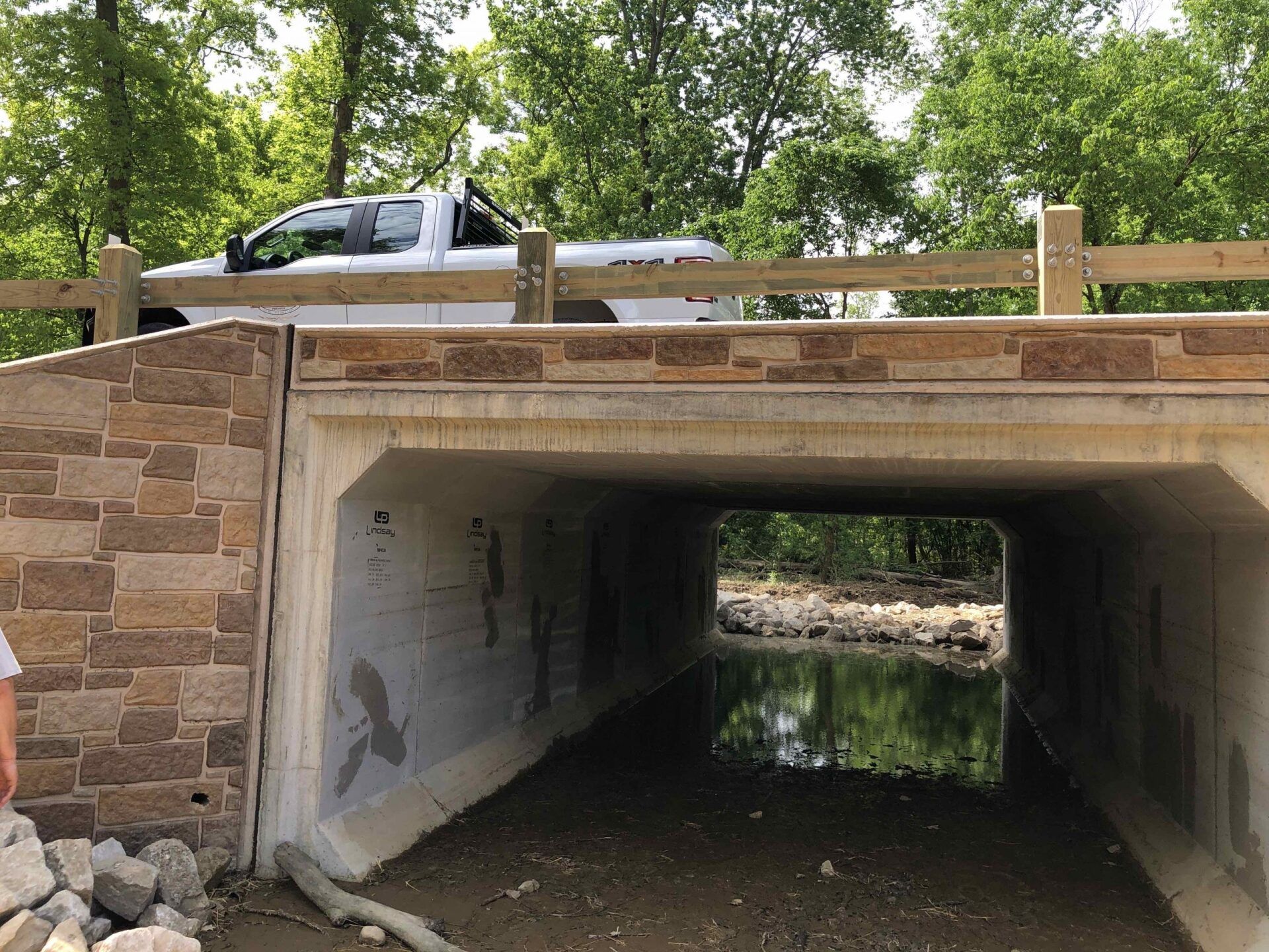Bridge over a creek. Stone sides, concrete top, and wooden railing. A white pickup truck is above the bridge.
