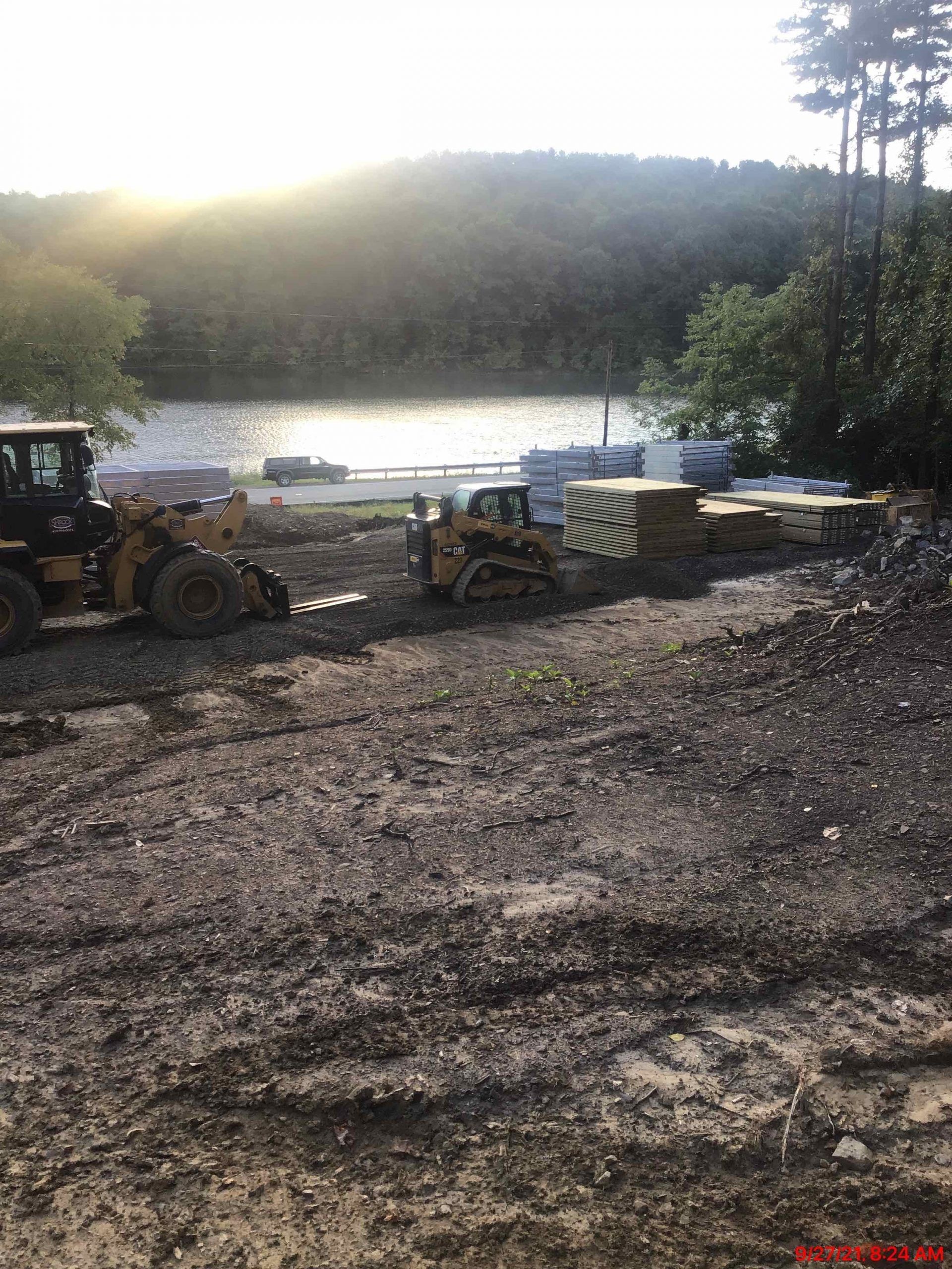 Construction site near water; bulldozer and equipment. Muddy ground, trees, and sunlight.