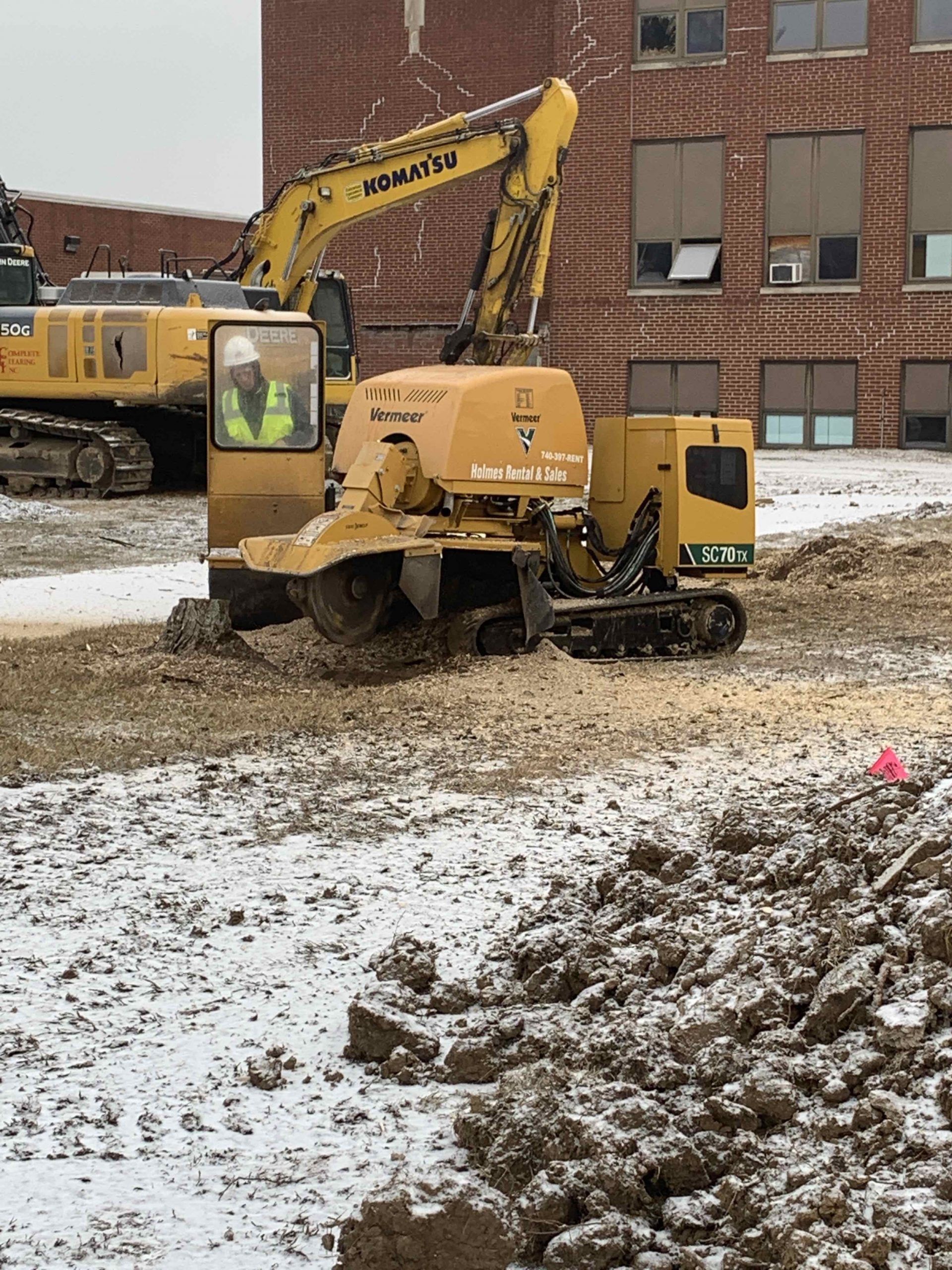 A tree stump grinder working in a snowy construction site near a brick building.