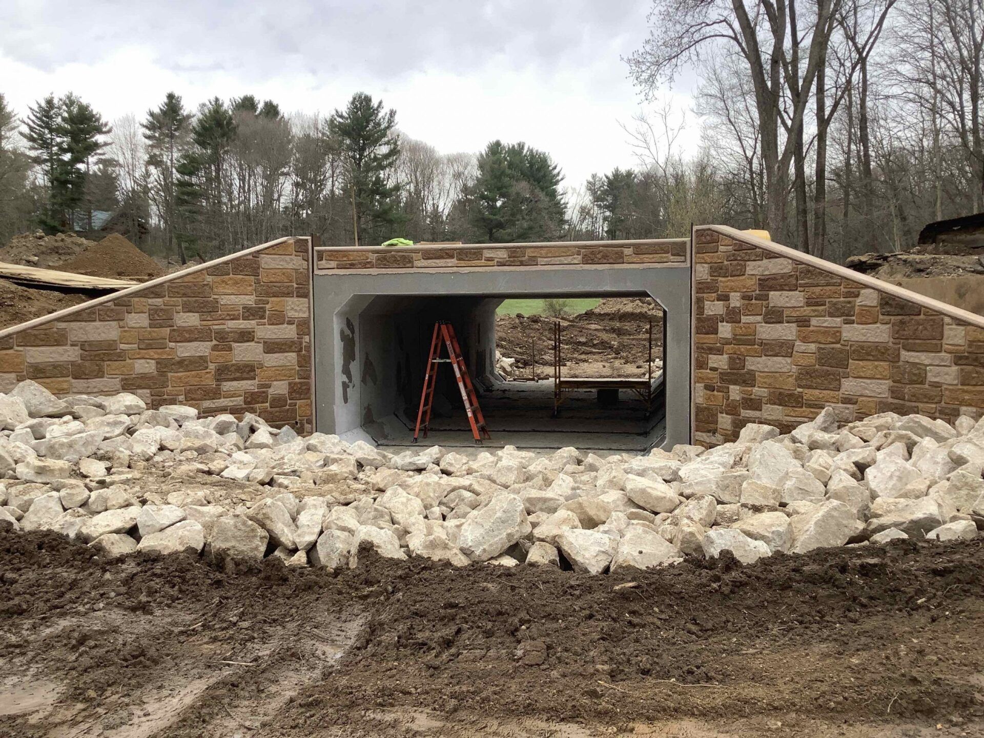 Stone-faced tunnel entrance under construction, with a ladder underneath. Surrounding the entrance is rock and dirt.