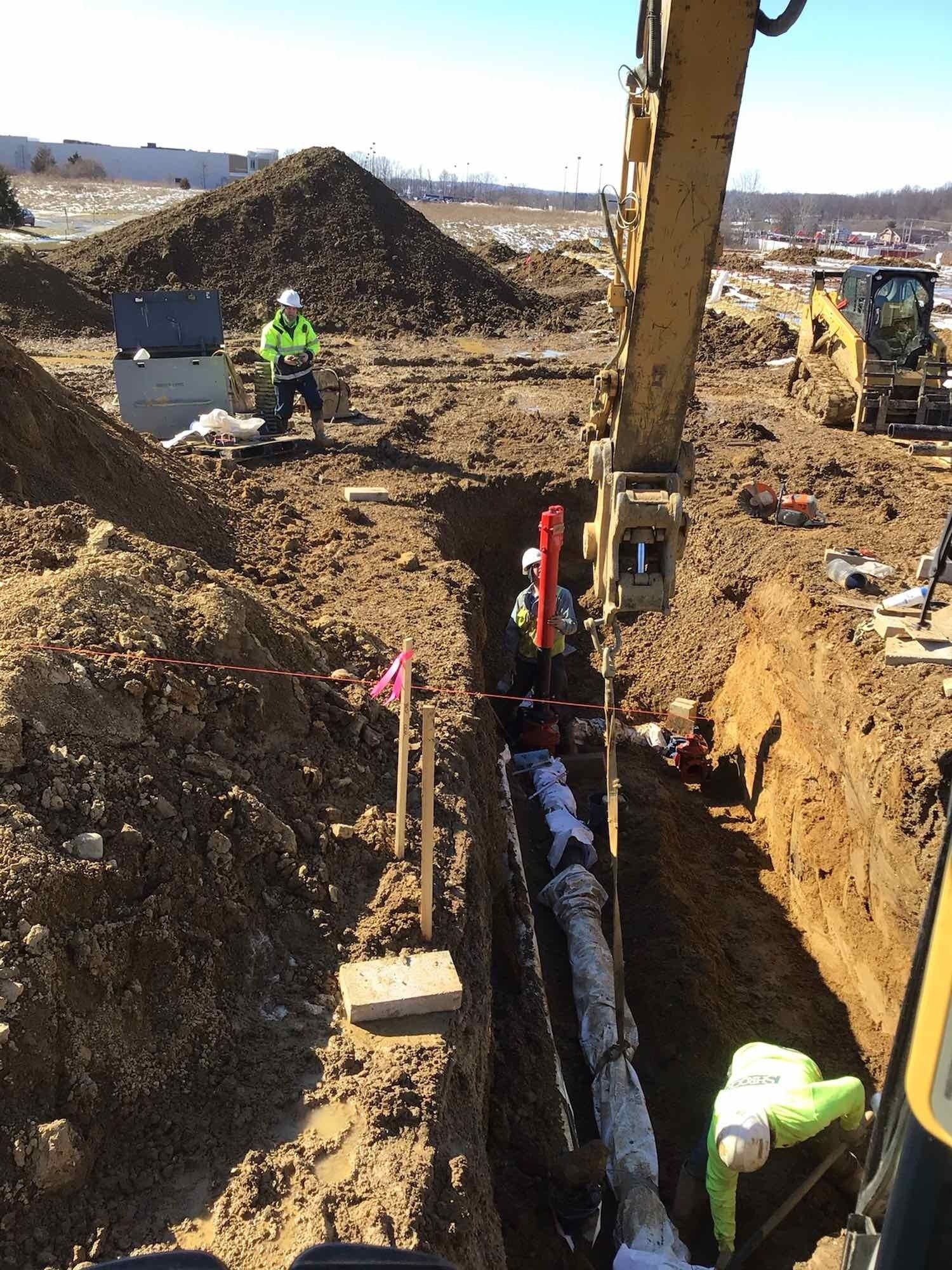 Construction workers installing a pipe in a trench; excavator overhead, safety vests visible; dirt pile in background.