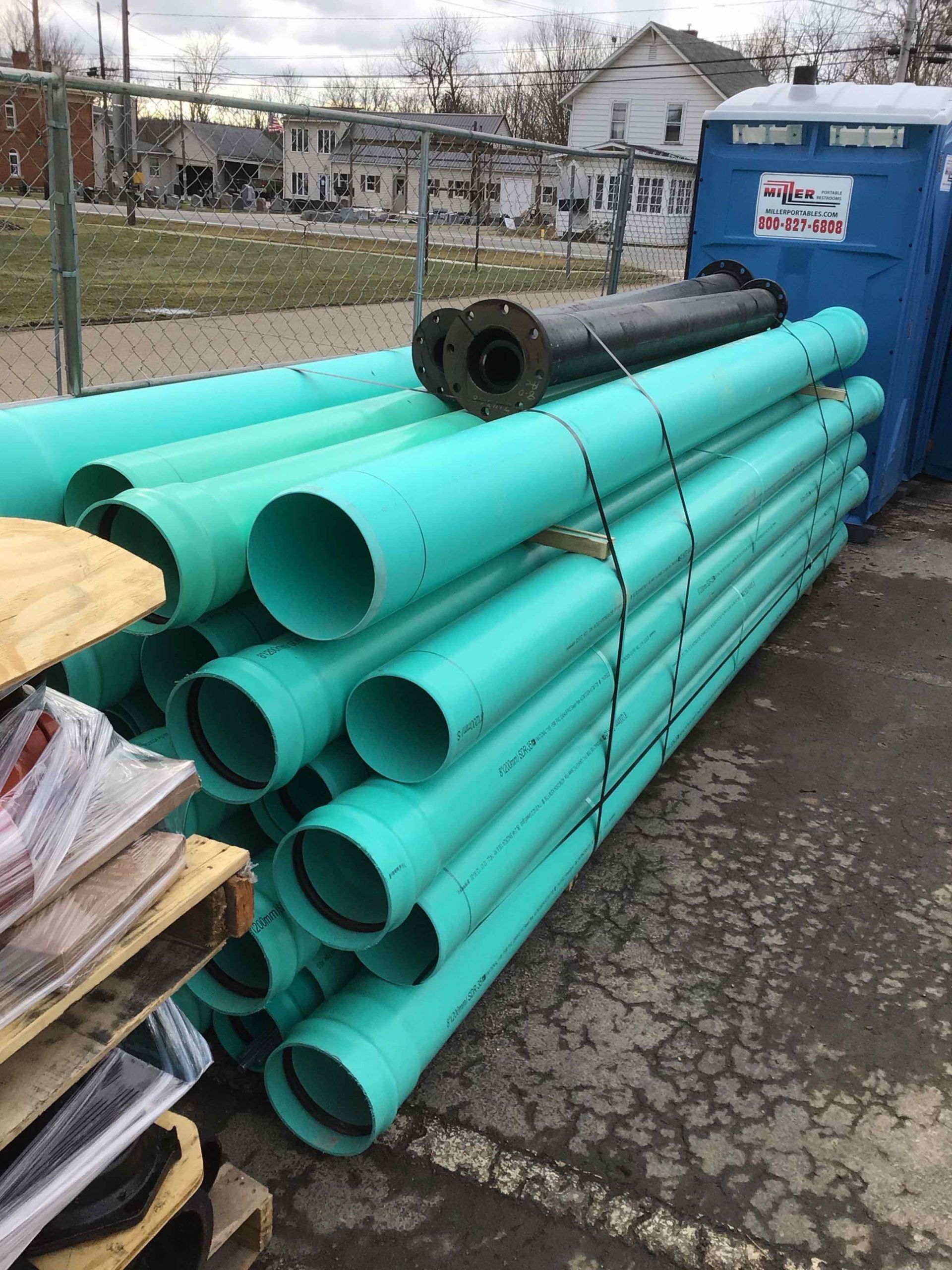 Green PVC pipes stacked outdoors near a blue portable toilet, possibly construction site.