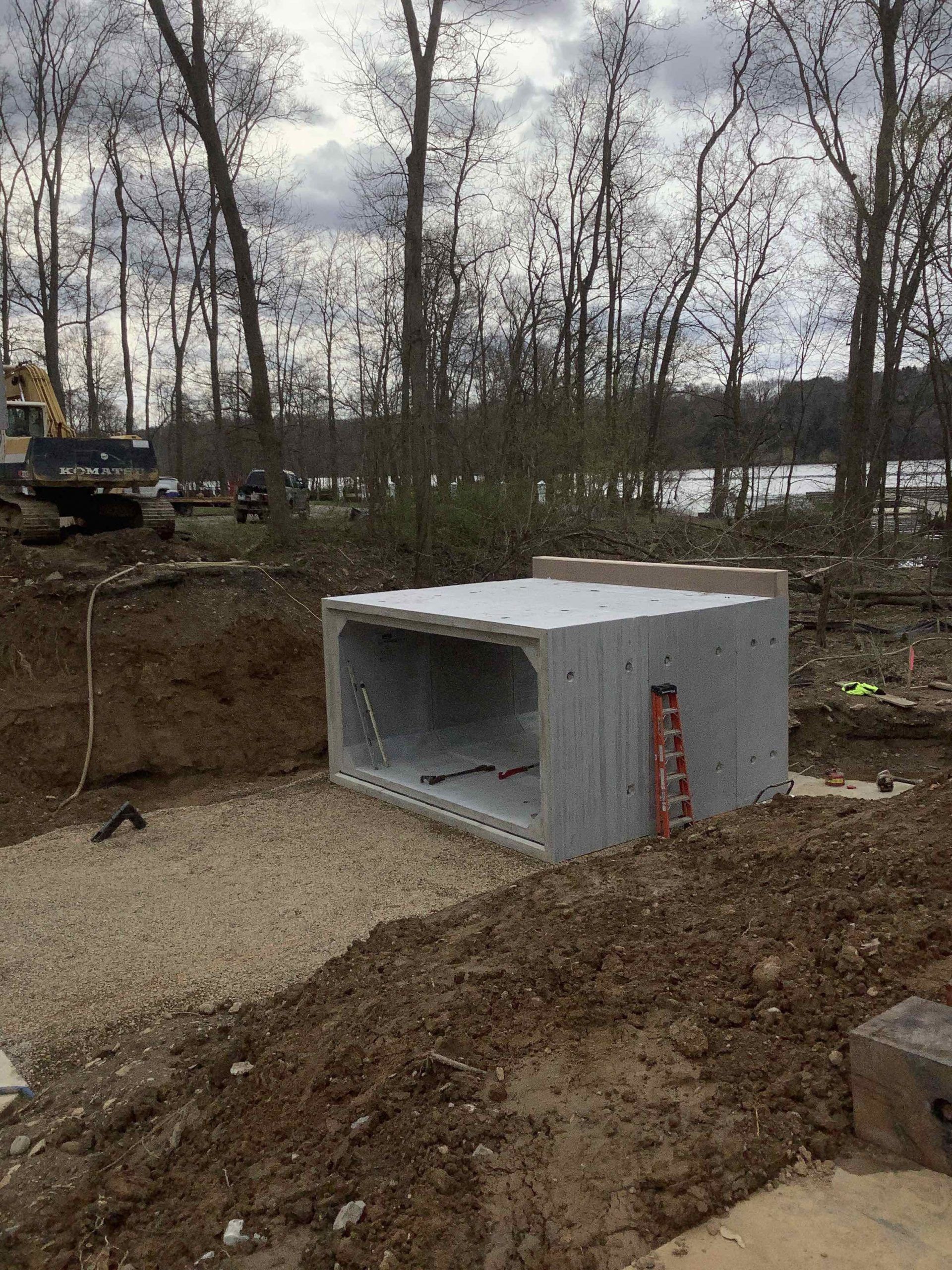 A large concrete box culvert sits in a construction site with trees and water in the background.
