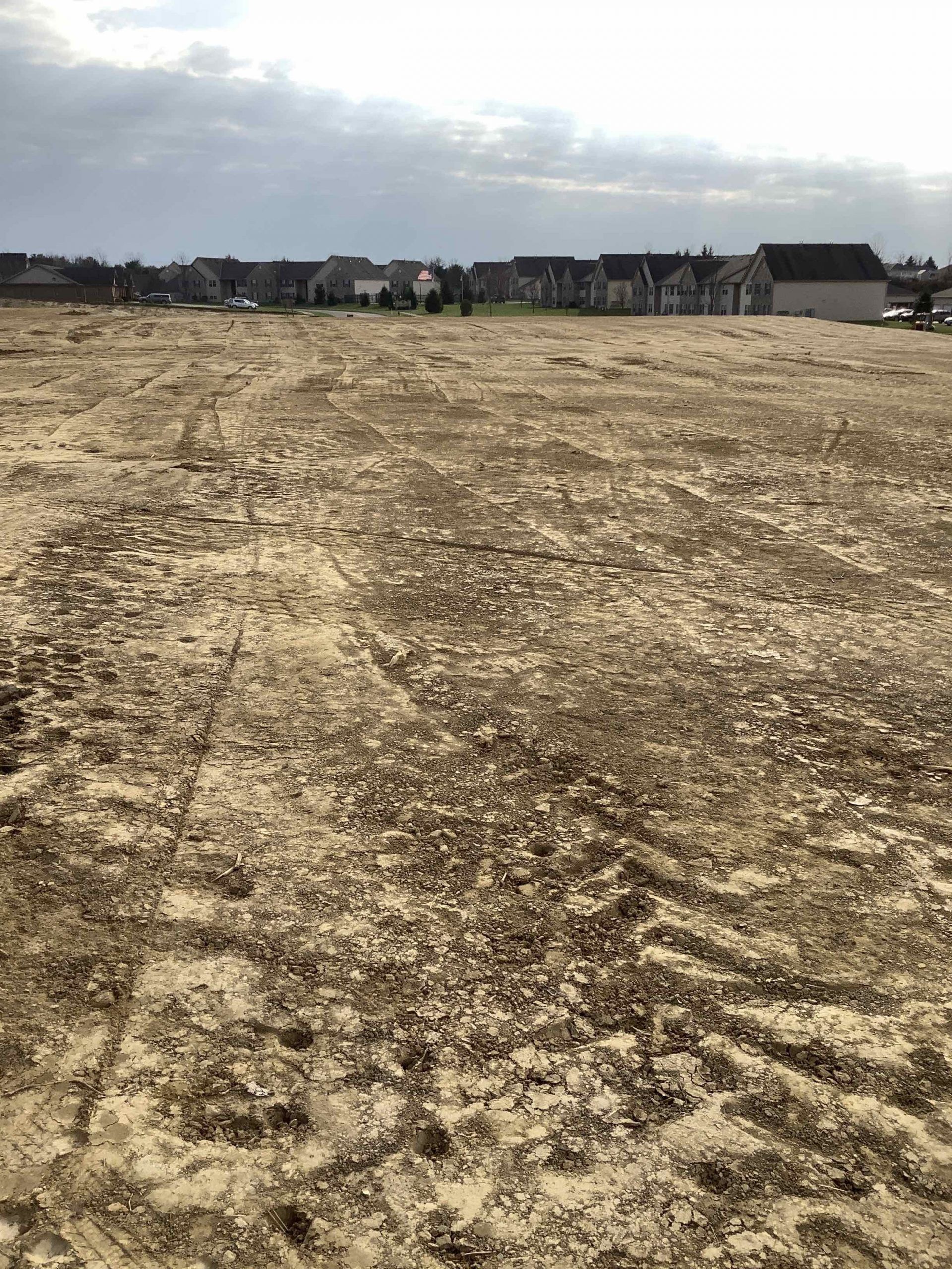 Cleared dirt field with tire tracks, houses in background under cloudy sky.