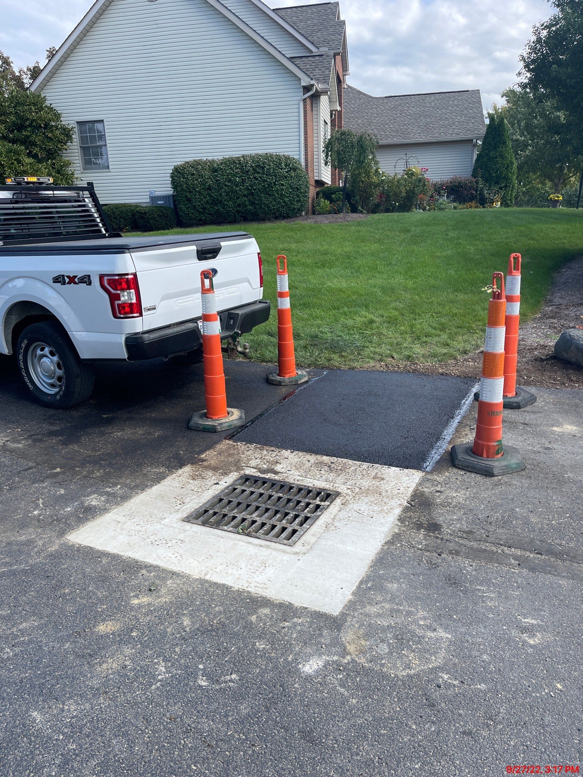 White truck next to a newly paved patch around a storm drain, marked by orange cones.