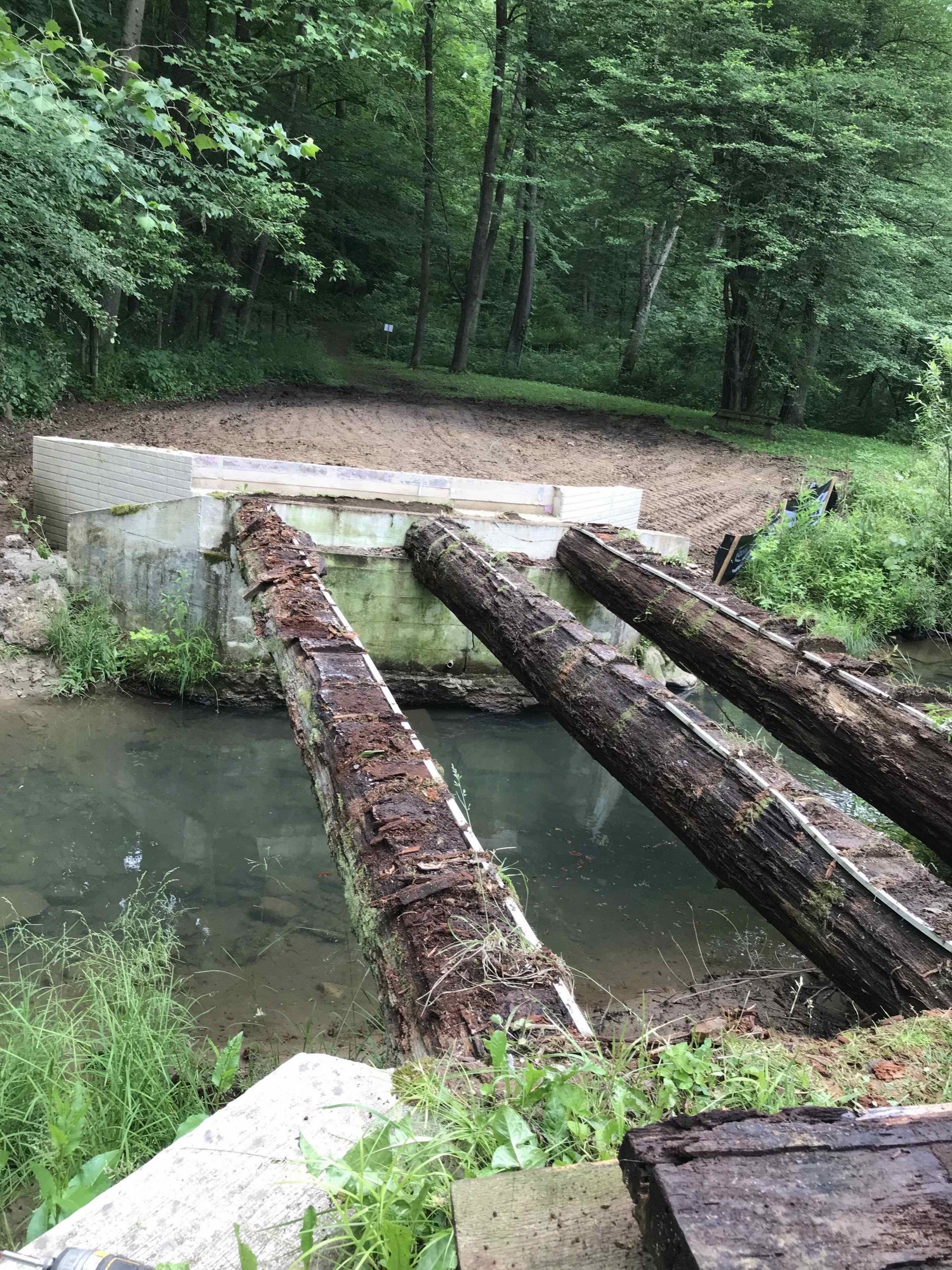 Wooden log bridge over a stream with concrete abutments; trees in the background.