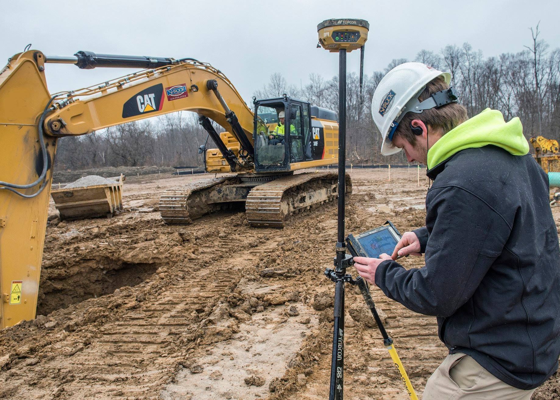 Construction worker using GPS device in muddy field near excavator.