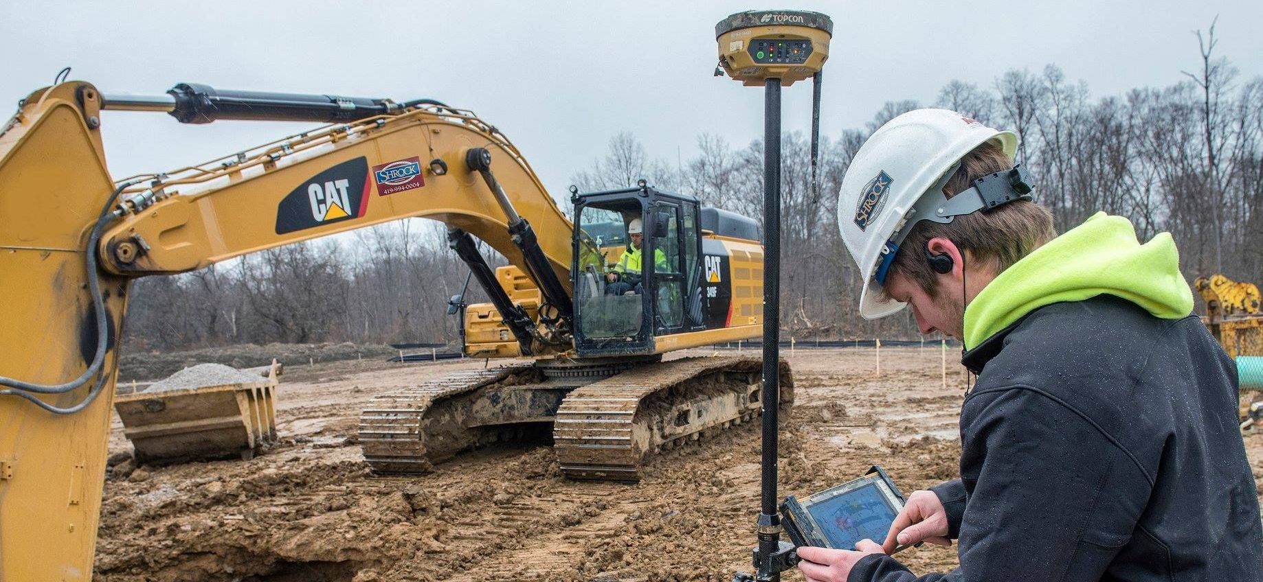 Construction worker with GPS device near an excavator on a muddy site. Cloudy day.