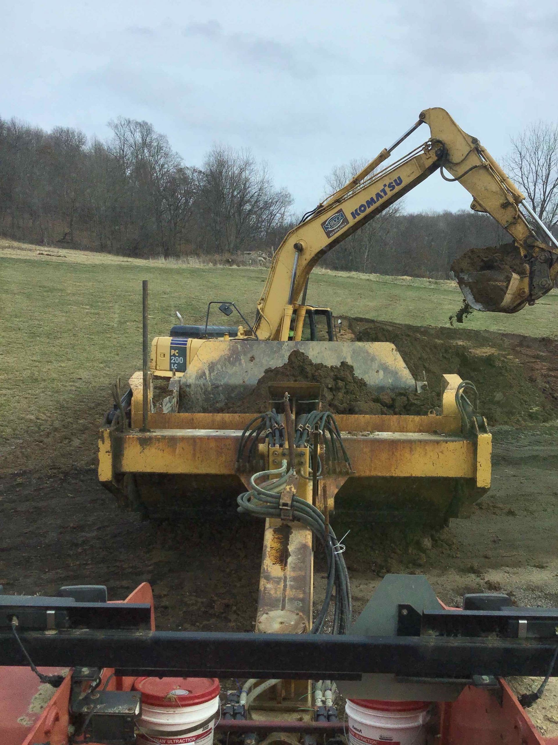 Yellow excavator dumping soil onto a scraper in a muddy field. Overcast day.