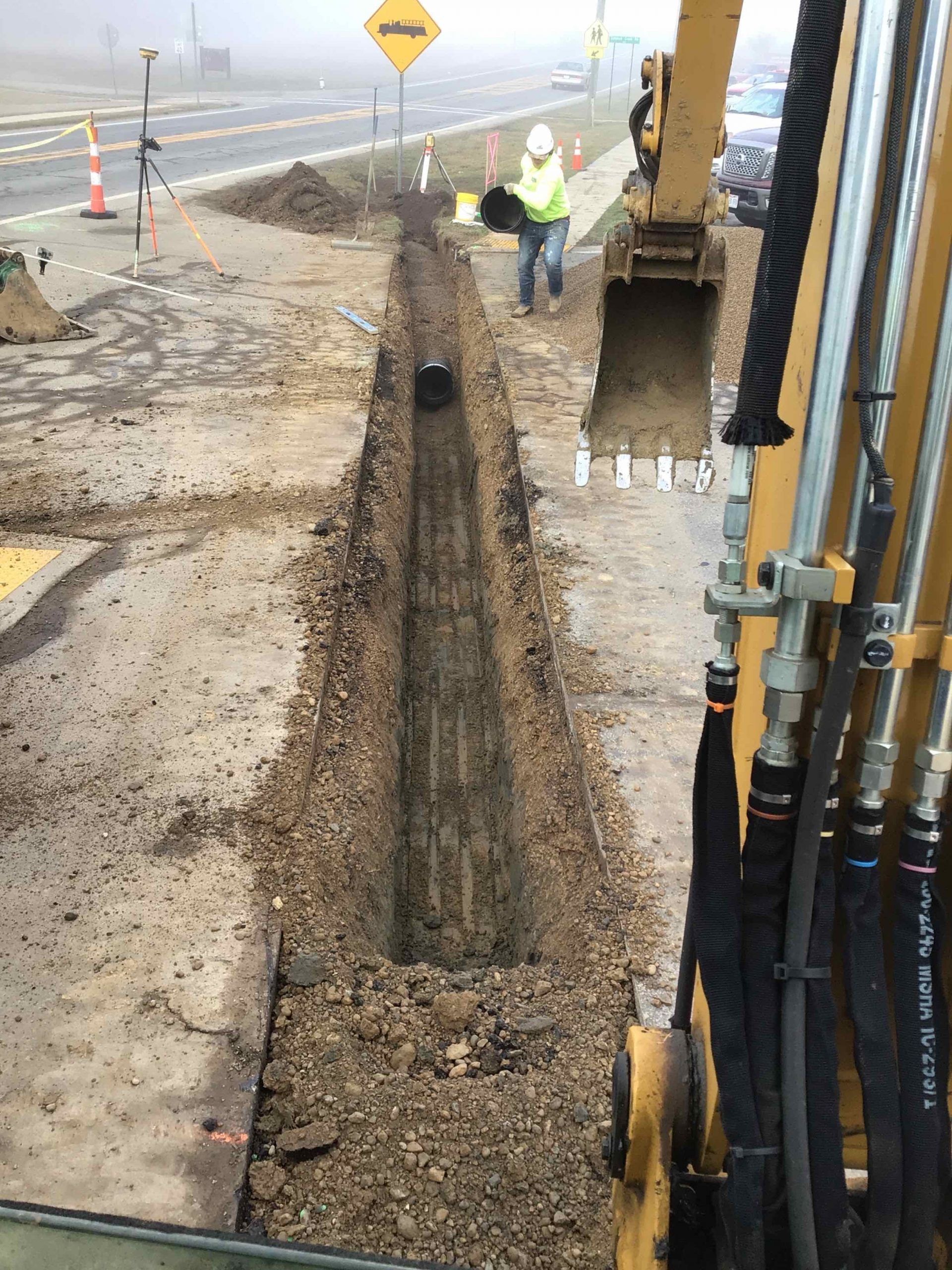 Construction site: Excavator digging a trench alongside a road, worker in high-vis vest.
