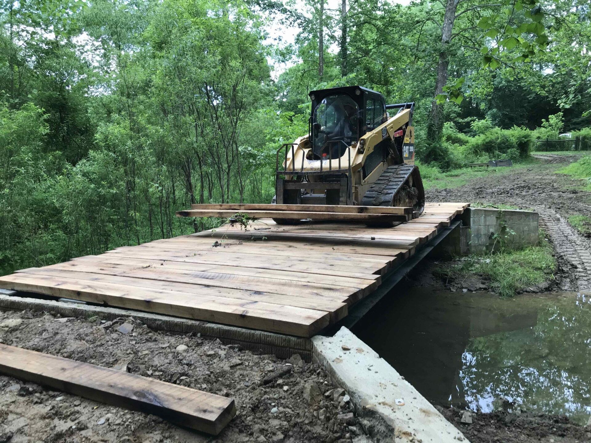 Skid steer loader on a wooden bridge crossing a muddy stream in a wooded area.