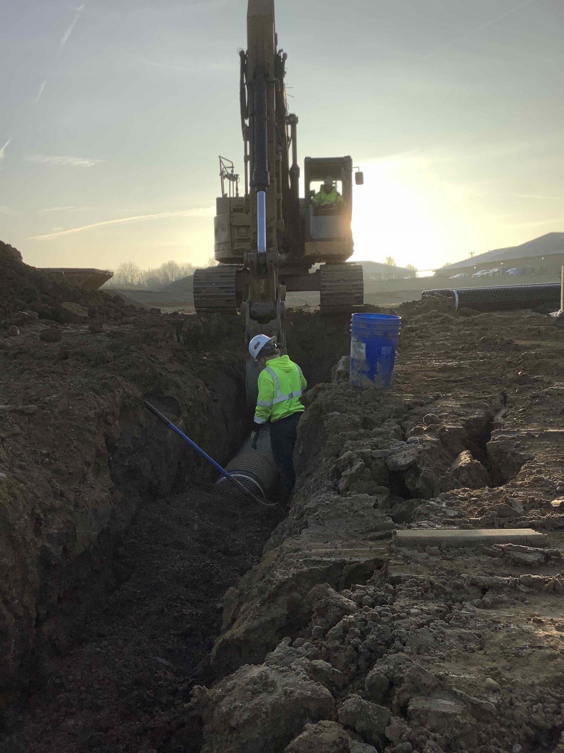 Construction worker in high-vis vest working in a trench with an excavator in the background.