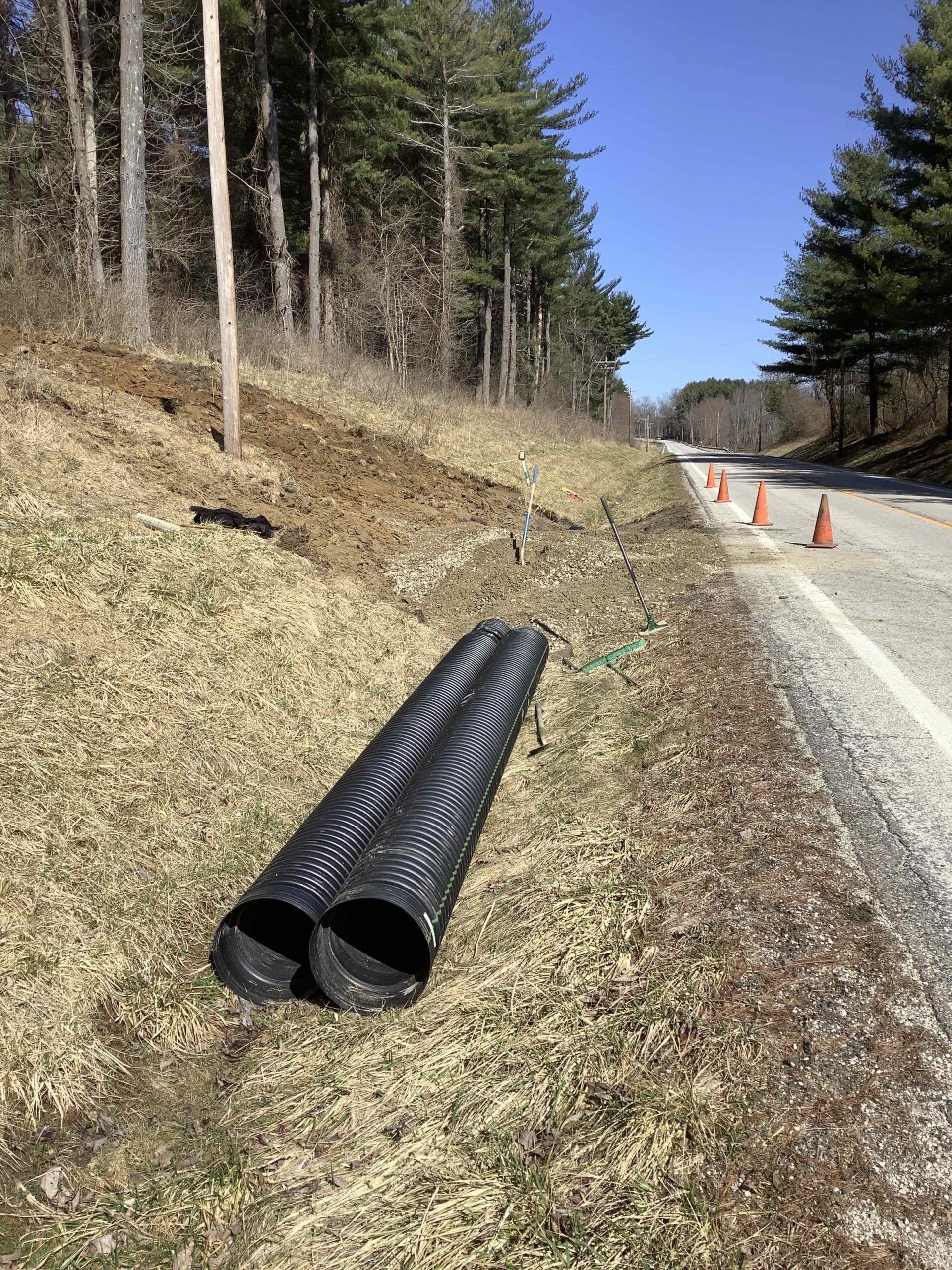 Black corrugated pipes lie near a roadside with orange cones and trees on a sunny day.