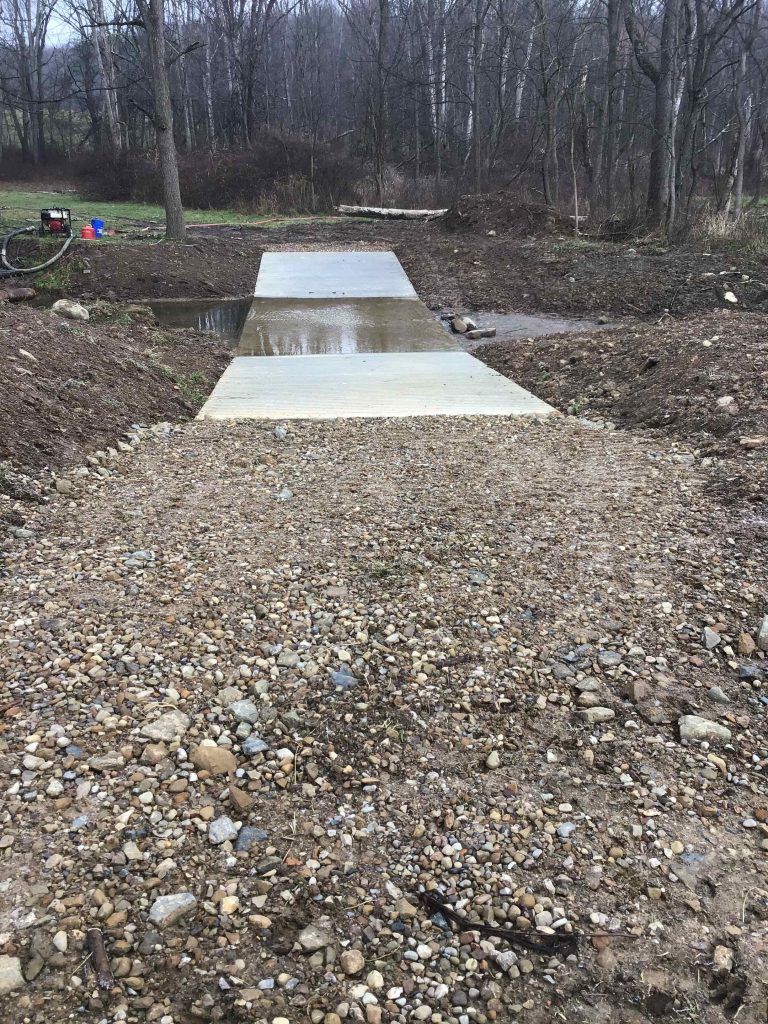 Gravel and concrete bridge over a shallow stream in a wooded area.