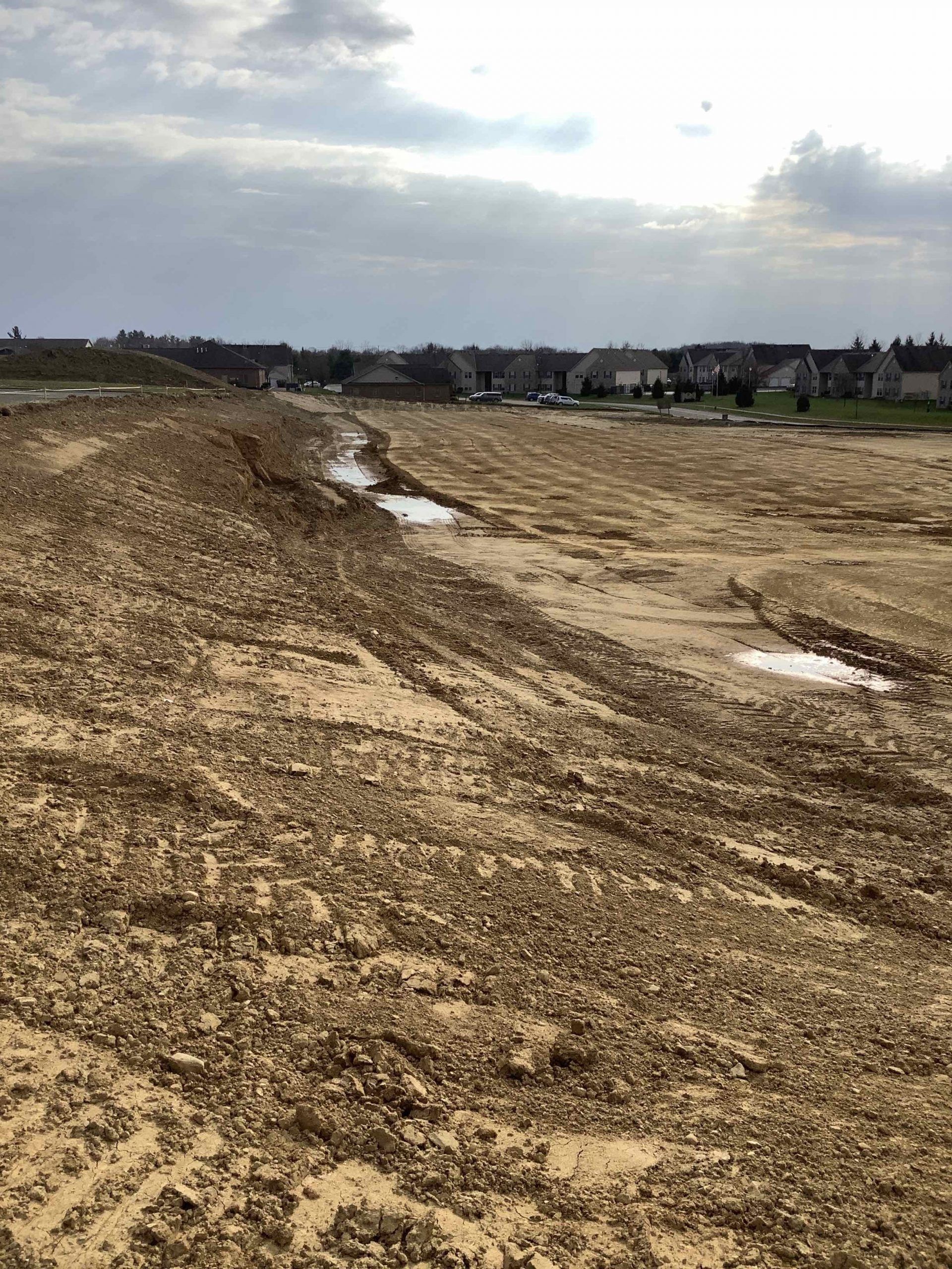 Dirt landscape with water puddles, construction in progress; houses in background under cloudy sky.