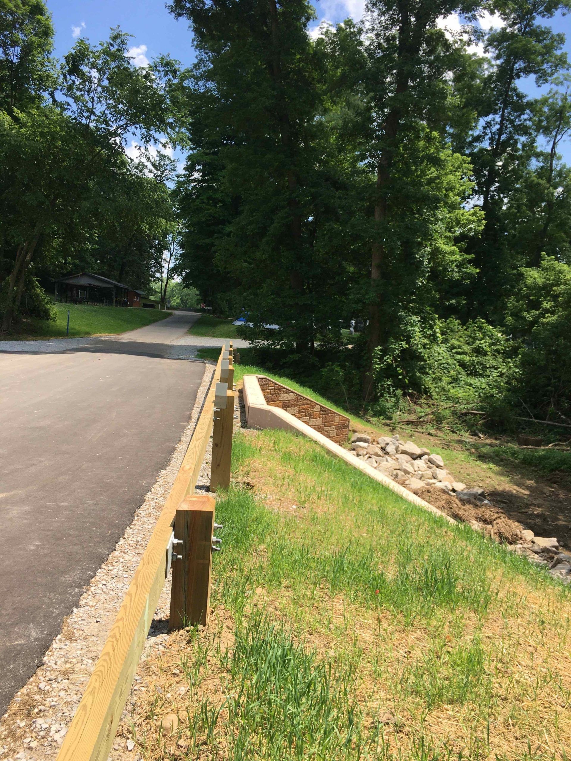 Paved path bordered by a wooden fence and grassy slope with trees in the background.