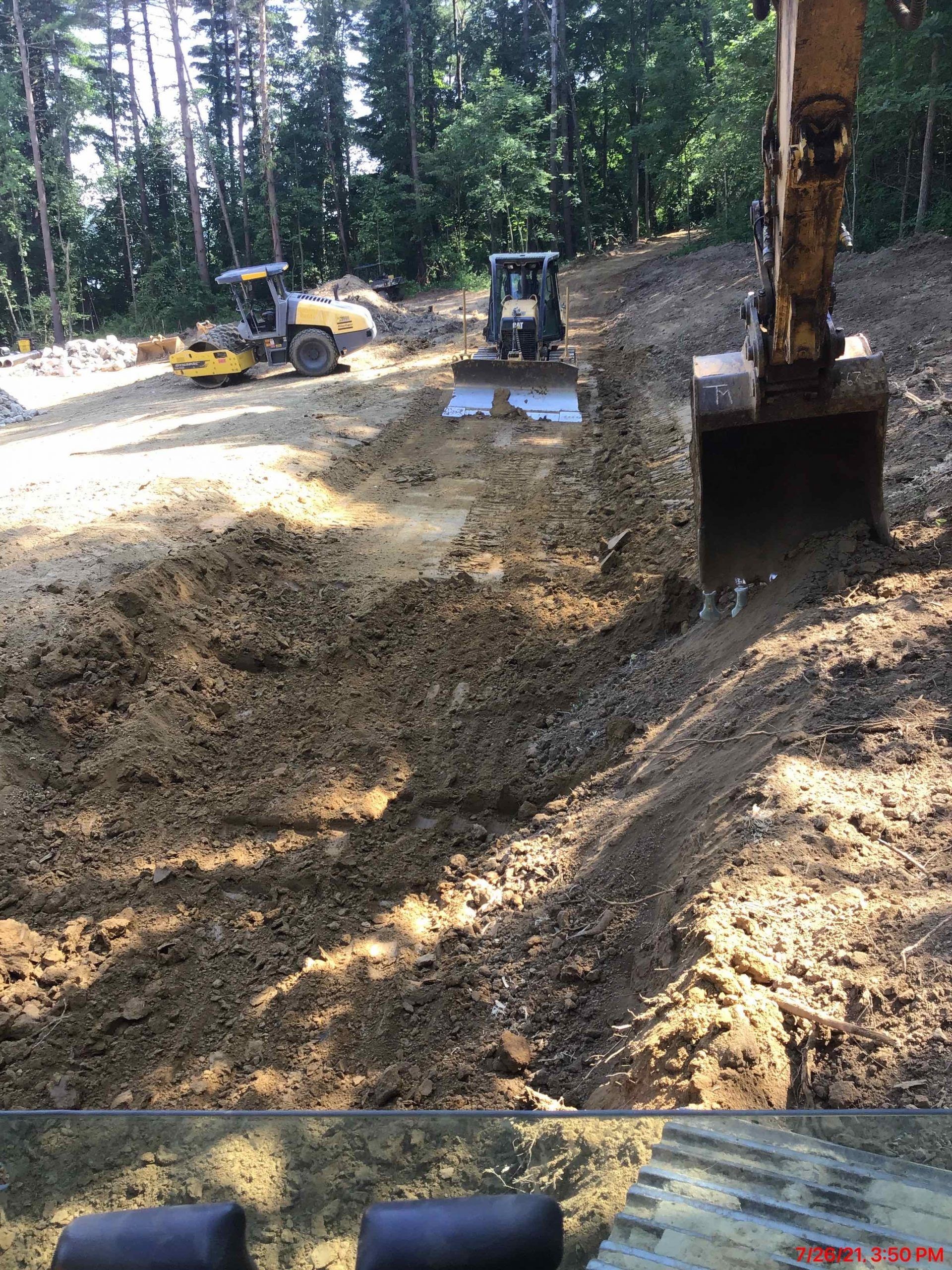 Construction site with excavator digging, two other machines in background; dirt, trees.