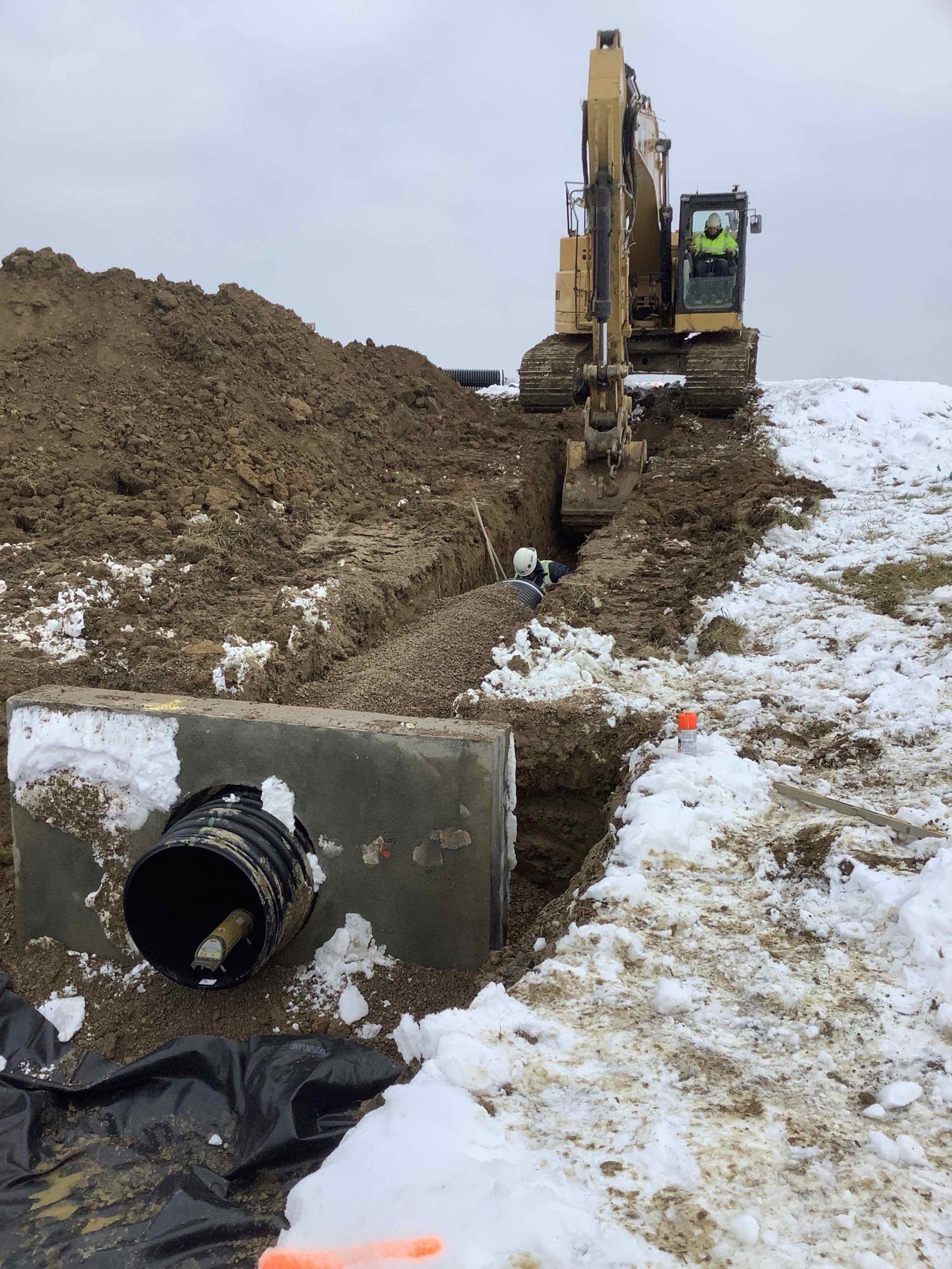 Excavator digging trench beside concrete box with pipe. Snow covers ground.