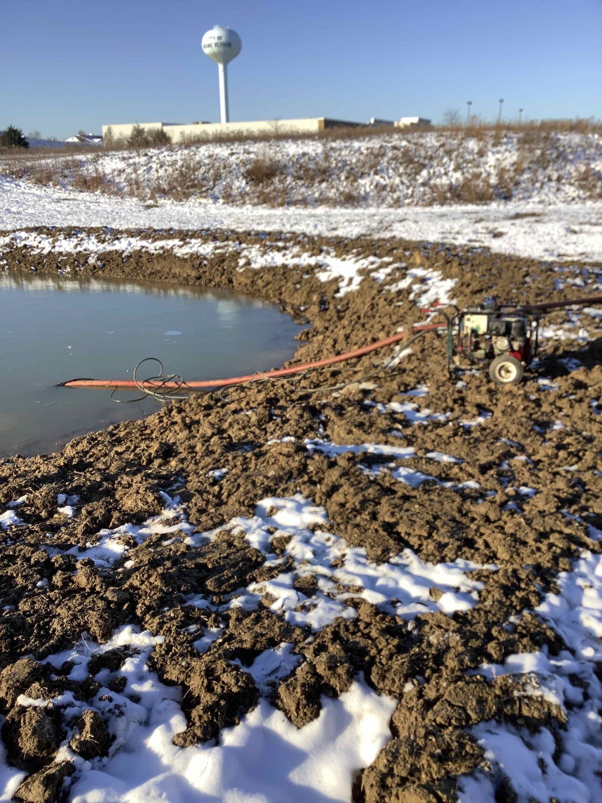 A pump and hose near a muddy pond in a snowy landscape, with a water tower in the background.