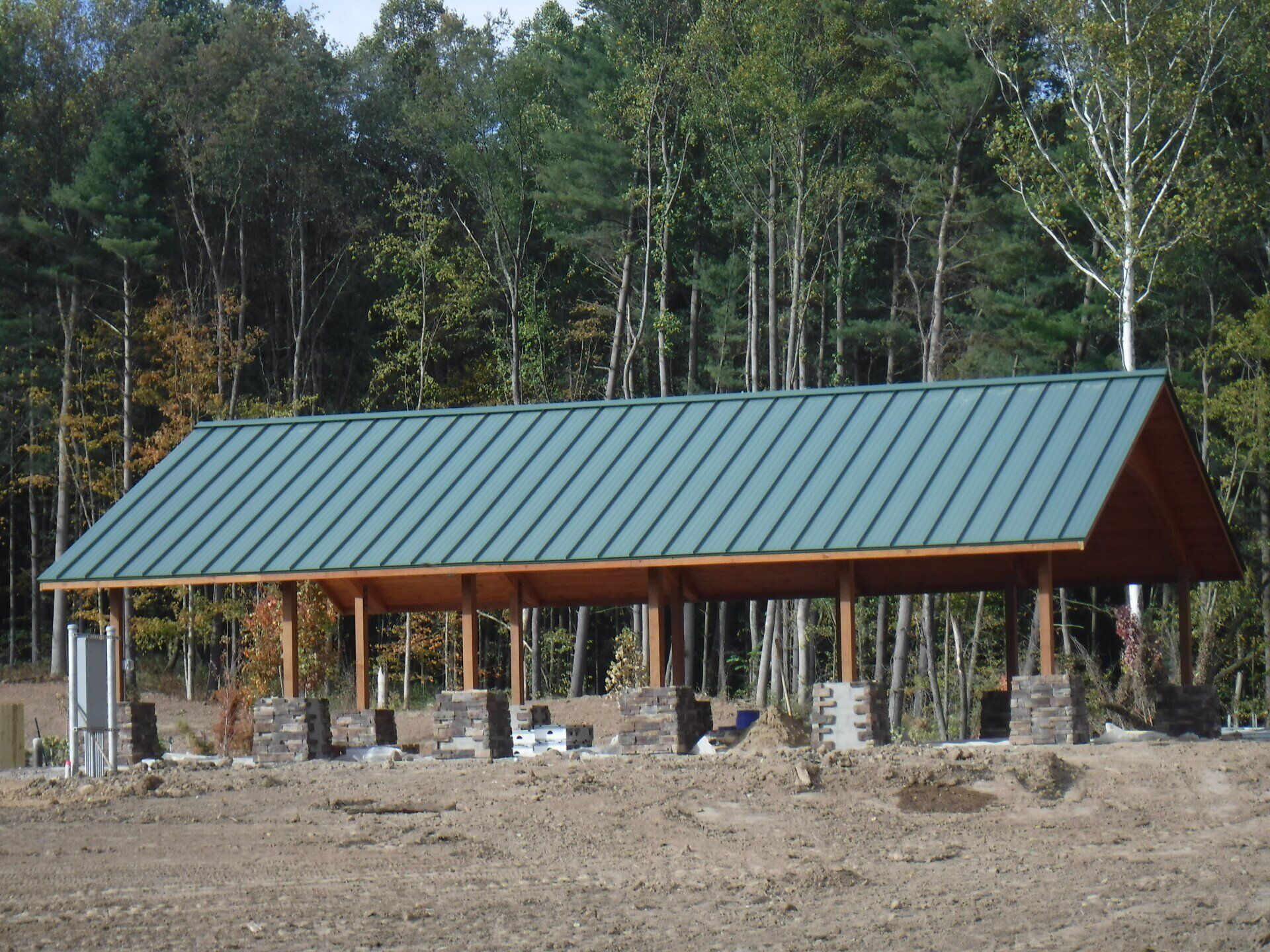 Green-roofed pavilion with wooden frame, supported by stone pillars, in a clearing near a forest.