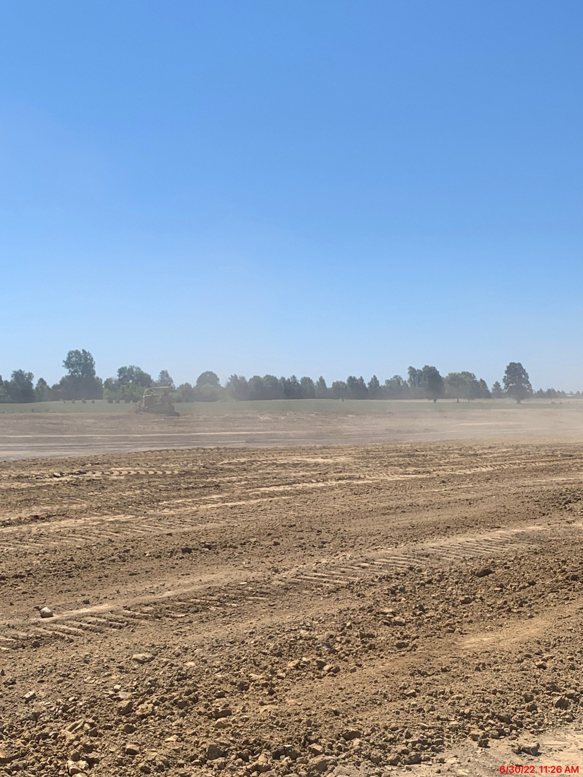 Field of dry, brown earth under a blue sky, with trees in the distance.