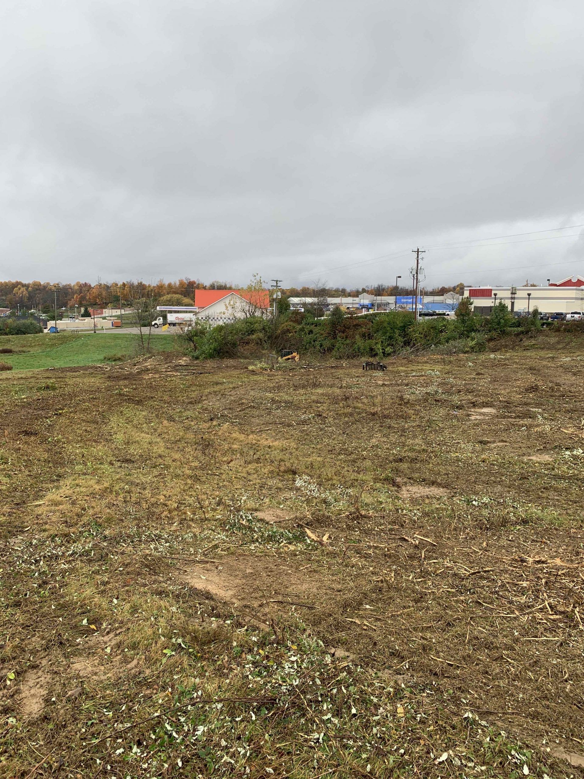 Overcast day, barren field in foreground, commercial buildings and trees in the distance.