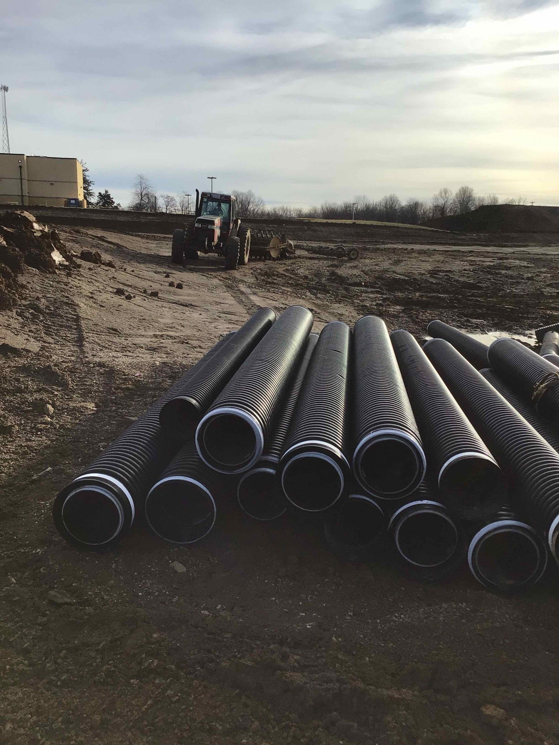 Black pipes with patterned ends stacked on dirt, tractor in background. Construction site.