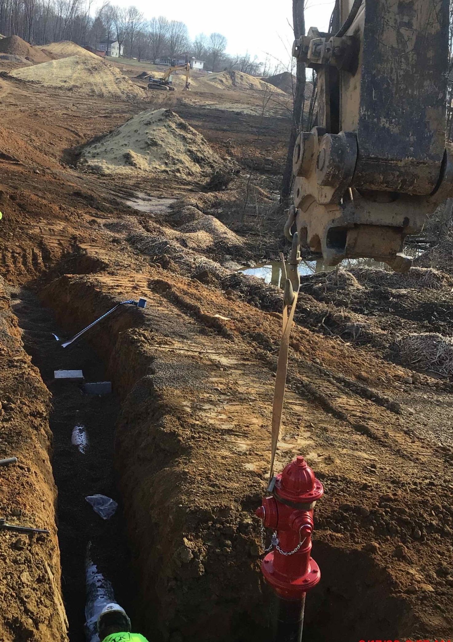 Construction site: Trench with pipes, red fire hydrant, backhoe arm. Brown earth, bright sunlight.