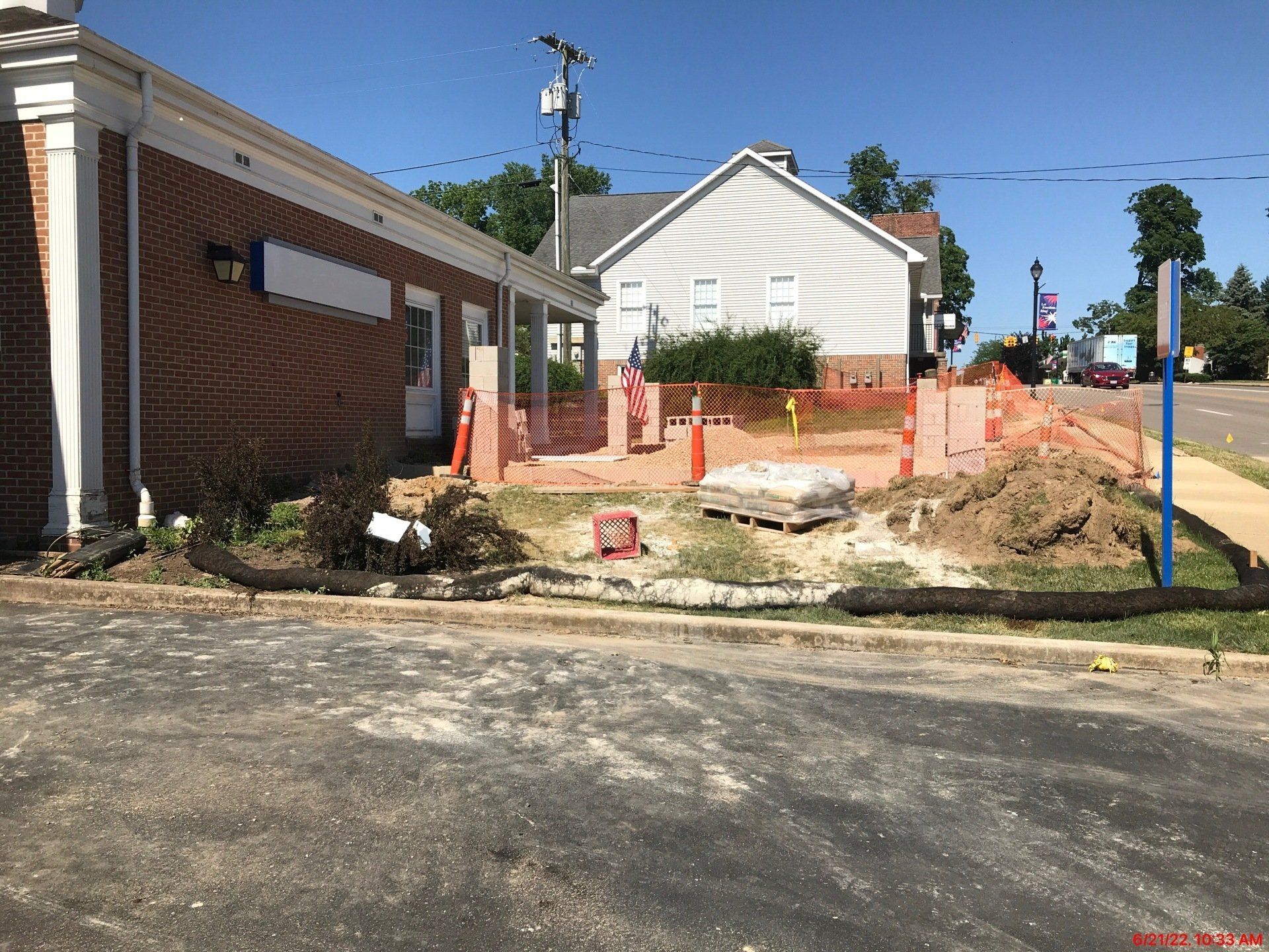 Construction site near a brick building and small white house, with orange safety barriers, dirt, and pavement.