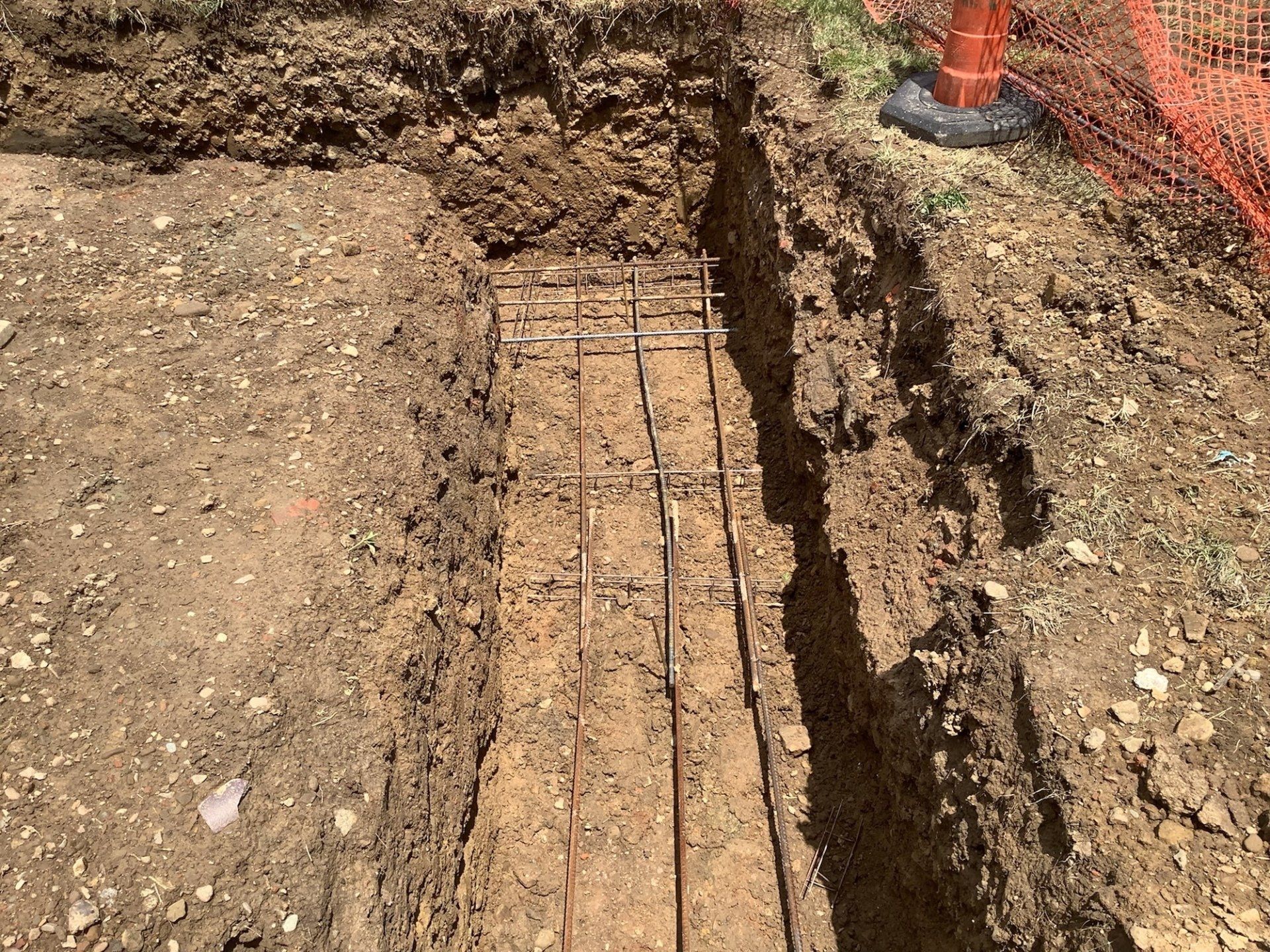 A rectangular trench dug in the dirt with a metal rebar grid inside, likely for a concrete foundation.