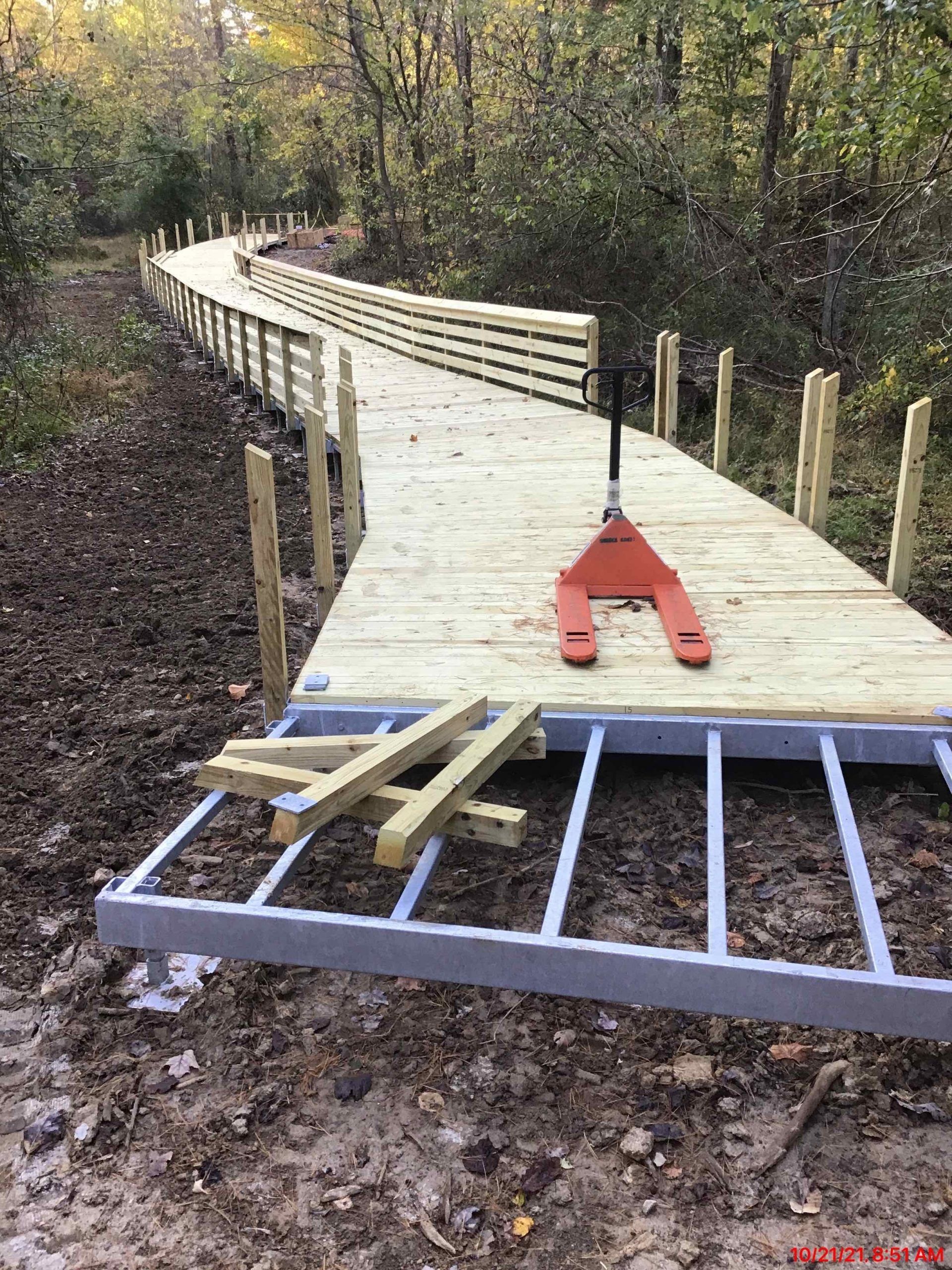 A wooden boardwalk under construction curves through a wooded area. An orange pallet jack sits on the boards.