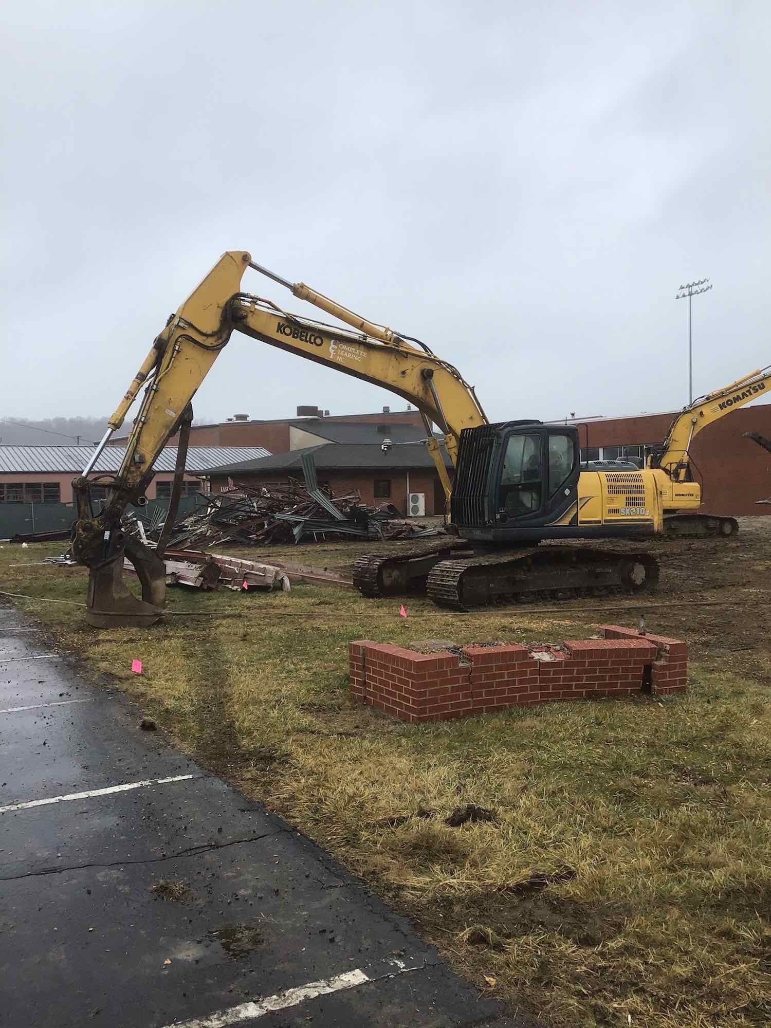 Yellow excavator demolishing a building, bricks in foreground, overcast day.