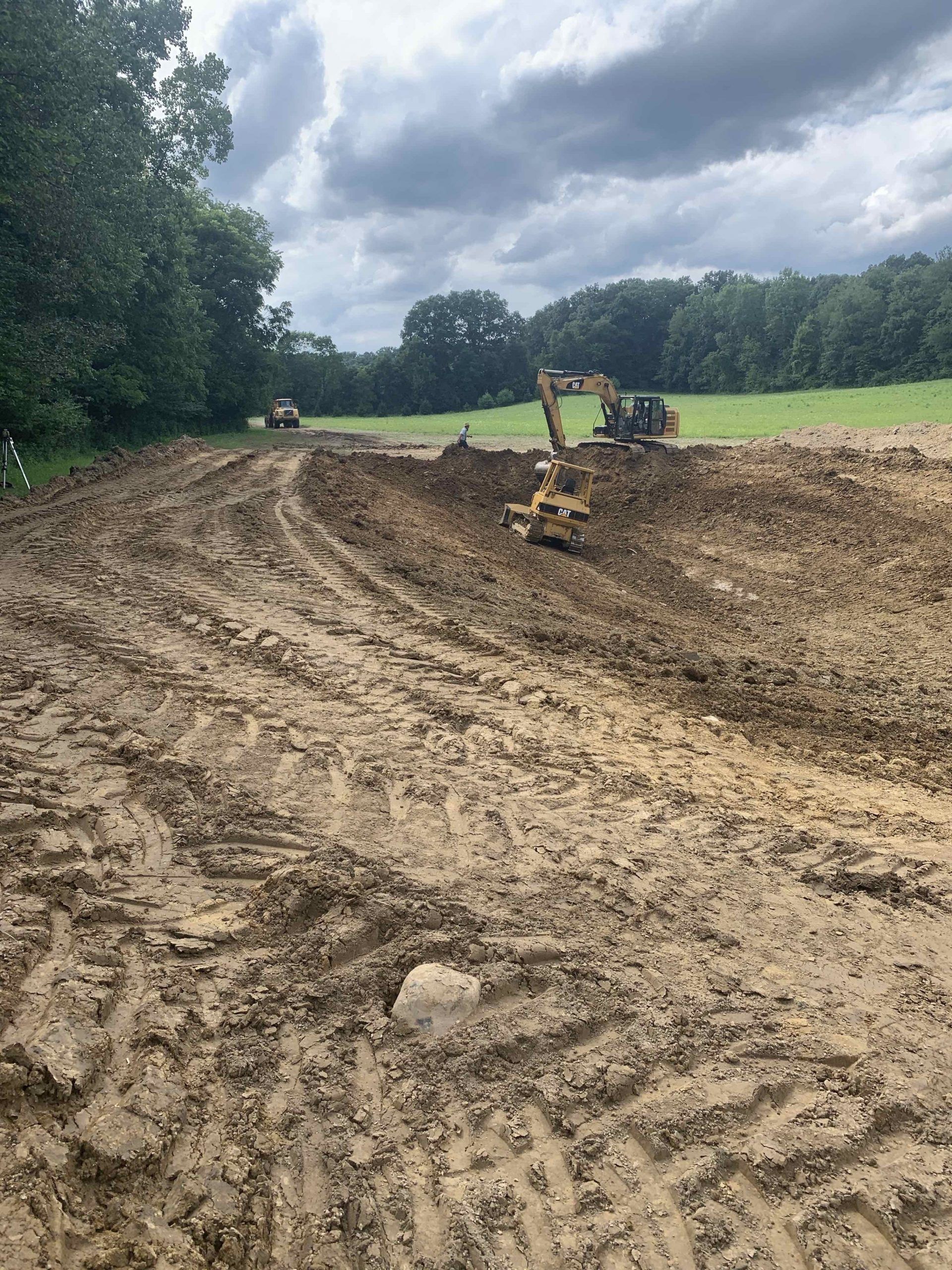 Muddy construction site with two excavators; a cloudy sky and treeline in the background.