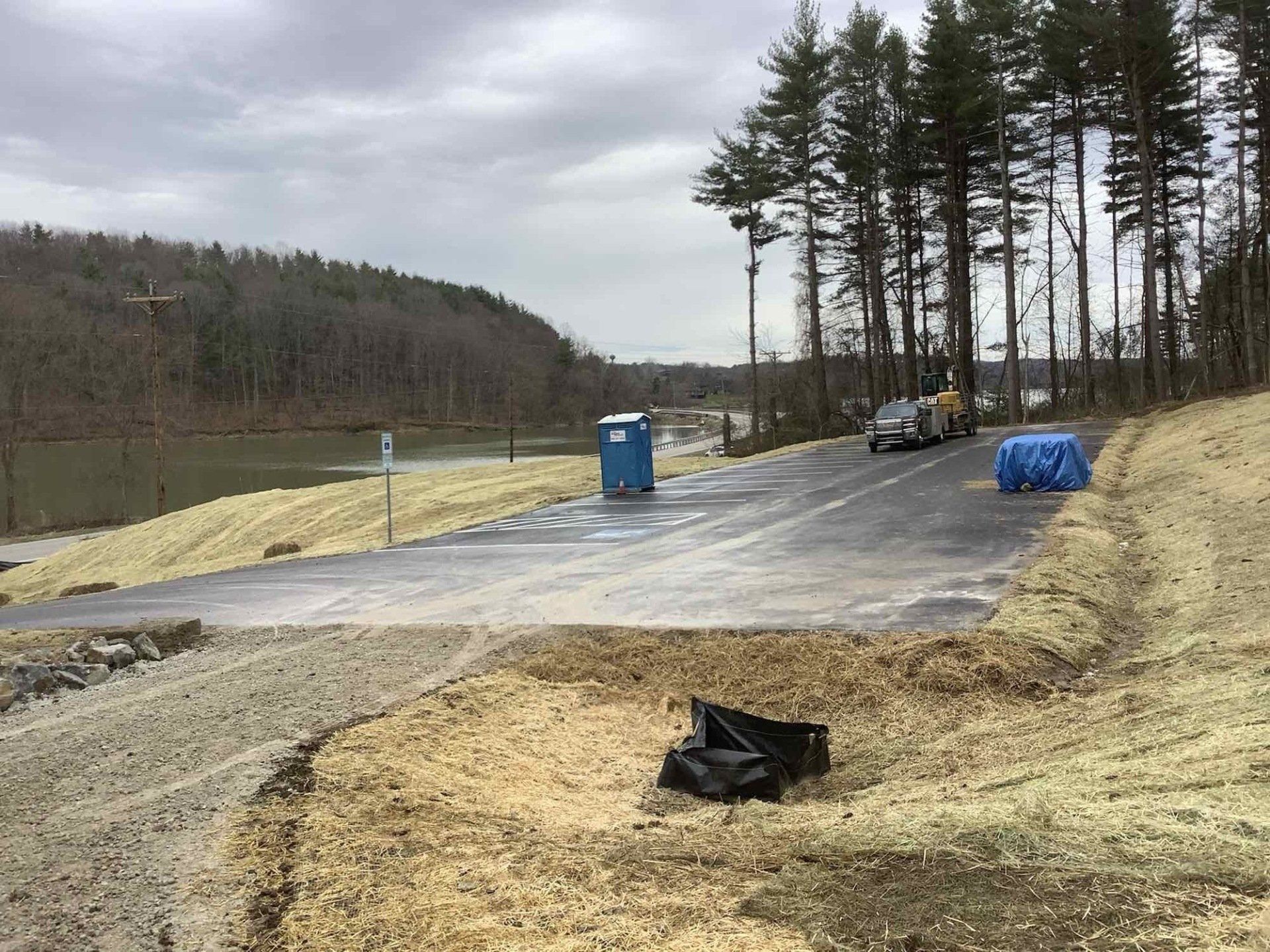Newly constructed parking area near a lake. Gravel edges, asphalt surface, portable toilet, and trees. Cloudy sky.