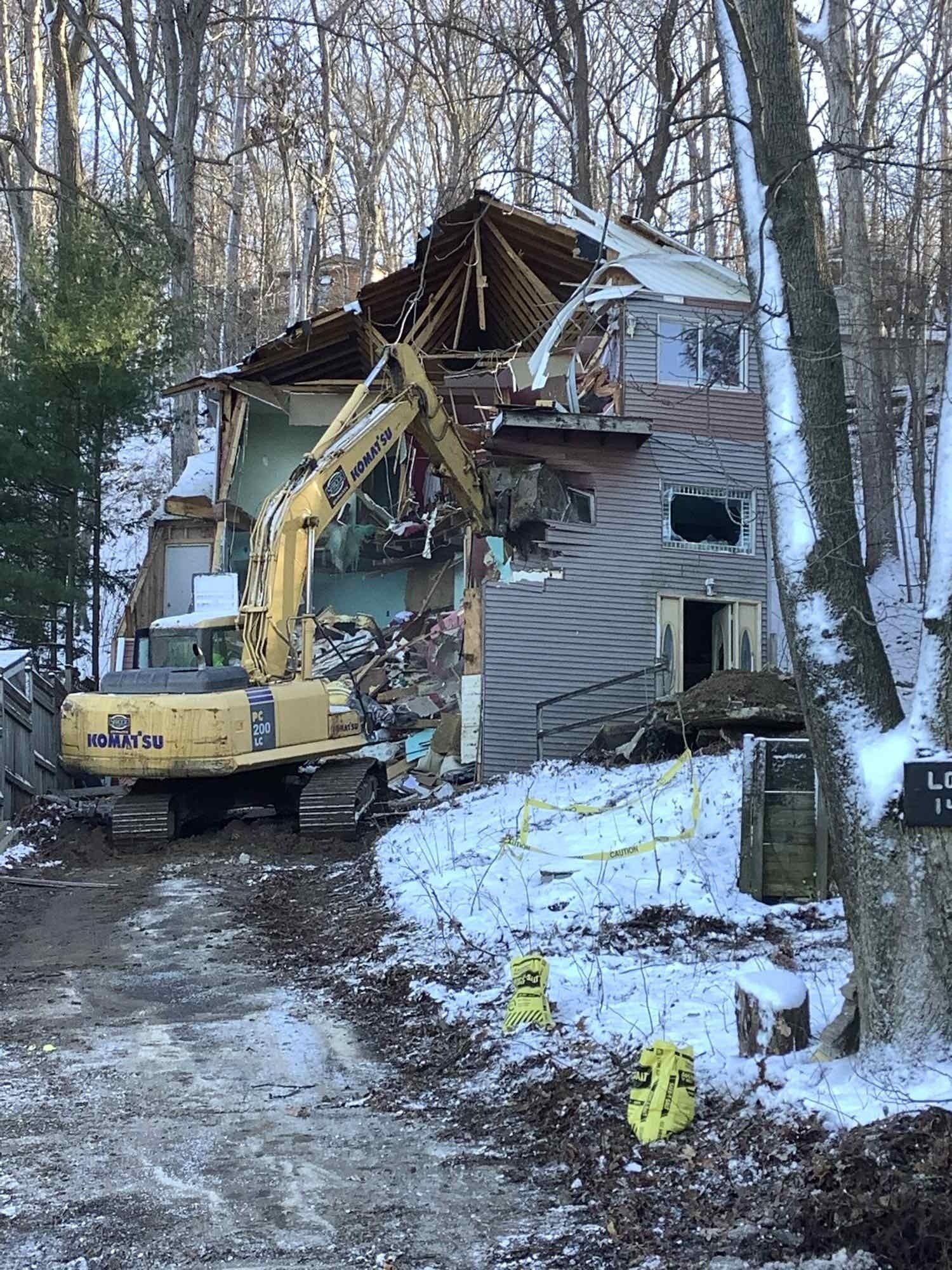 Demolition of a house in winter; an excavator tears apart the roof and walls, snow on the ground.