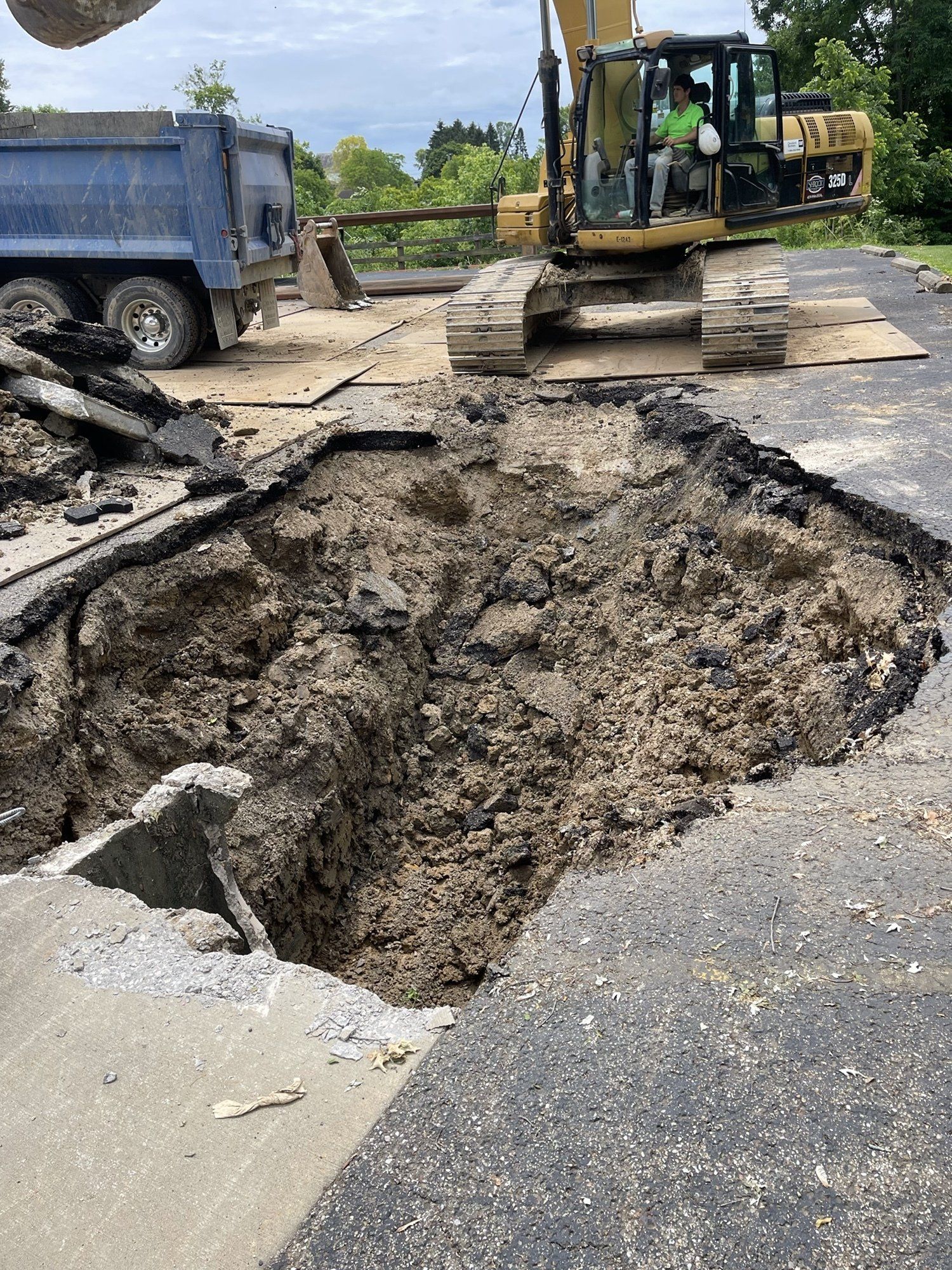 Excavator repairing a large hole in asphalt road; dump truck nearby. Cloudy day.