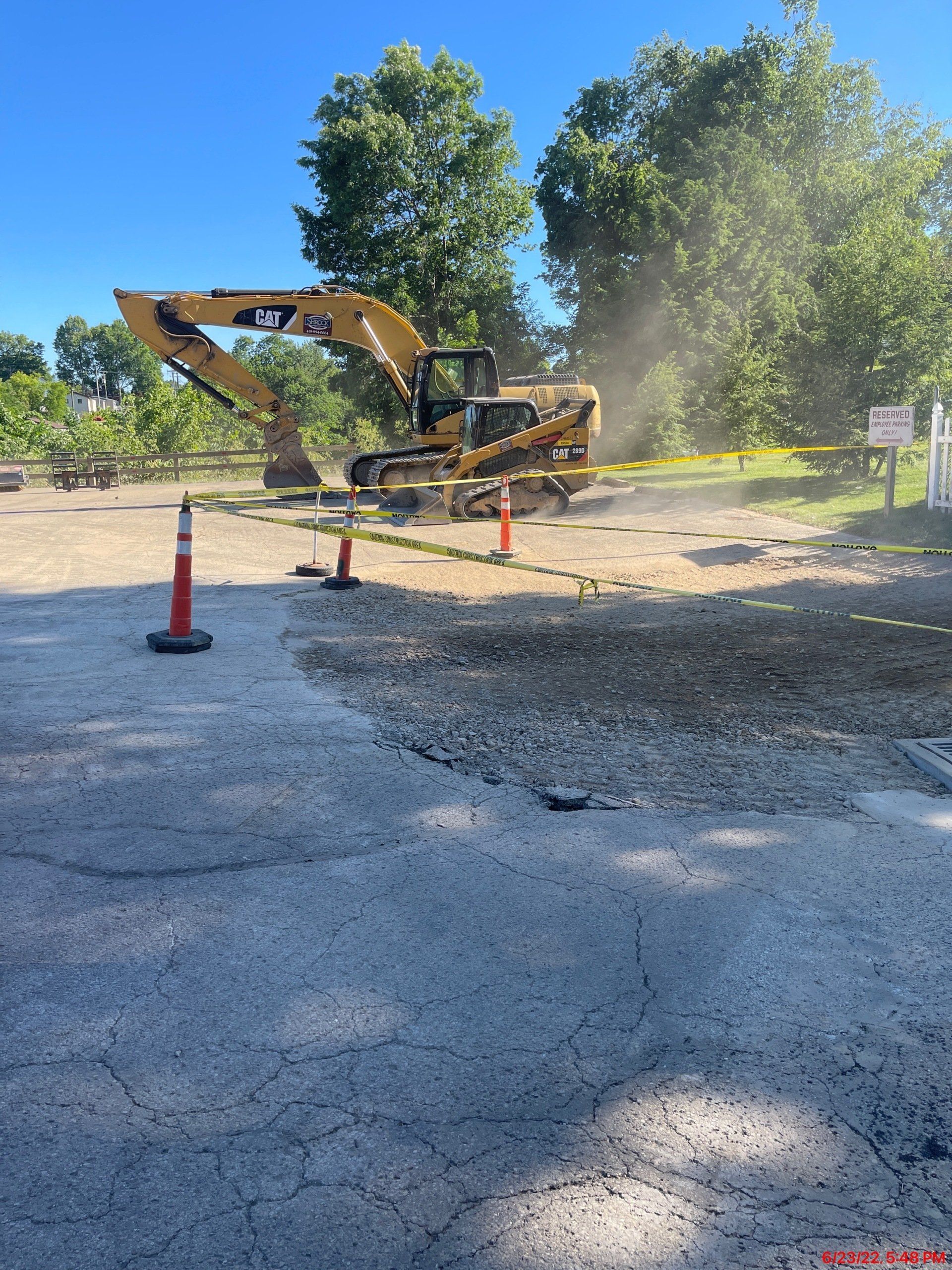 Yellow excavator on a gravel surface, possibly a parking lot. Safety cones and caution tape present. Trees in the background.
