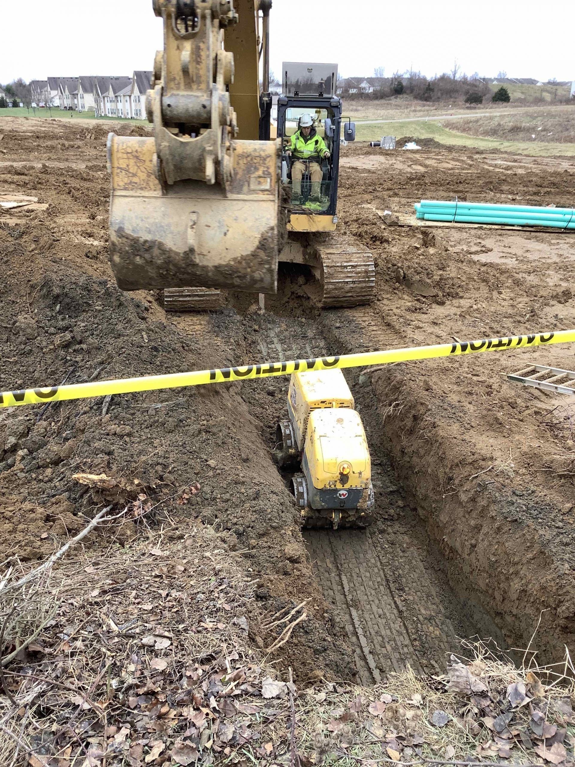 An excavator operator working at a construction site, with a trench and a compactor.