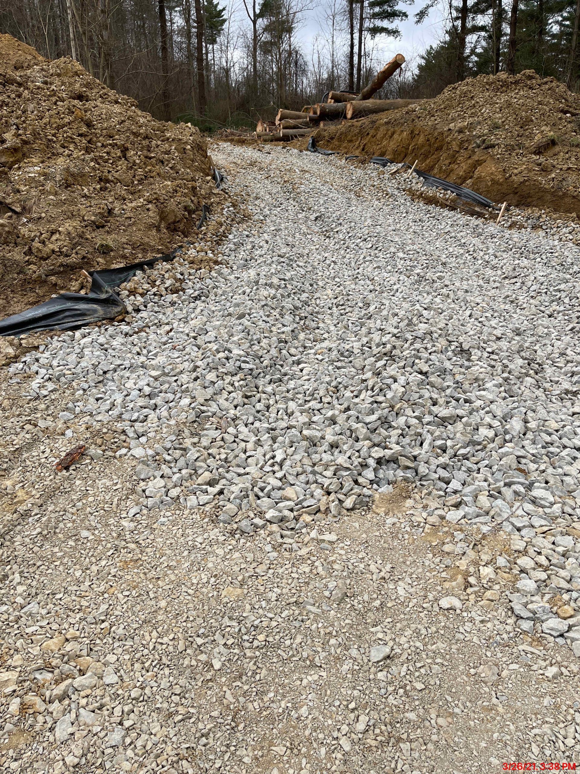 Gravel pathway flanked by dirt piles, leading through a forest.