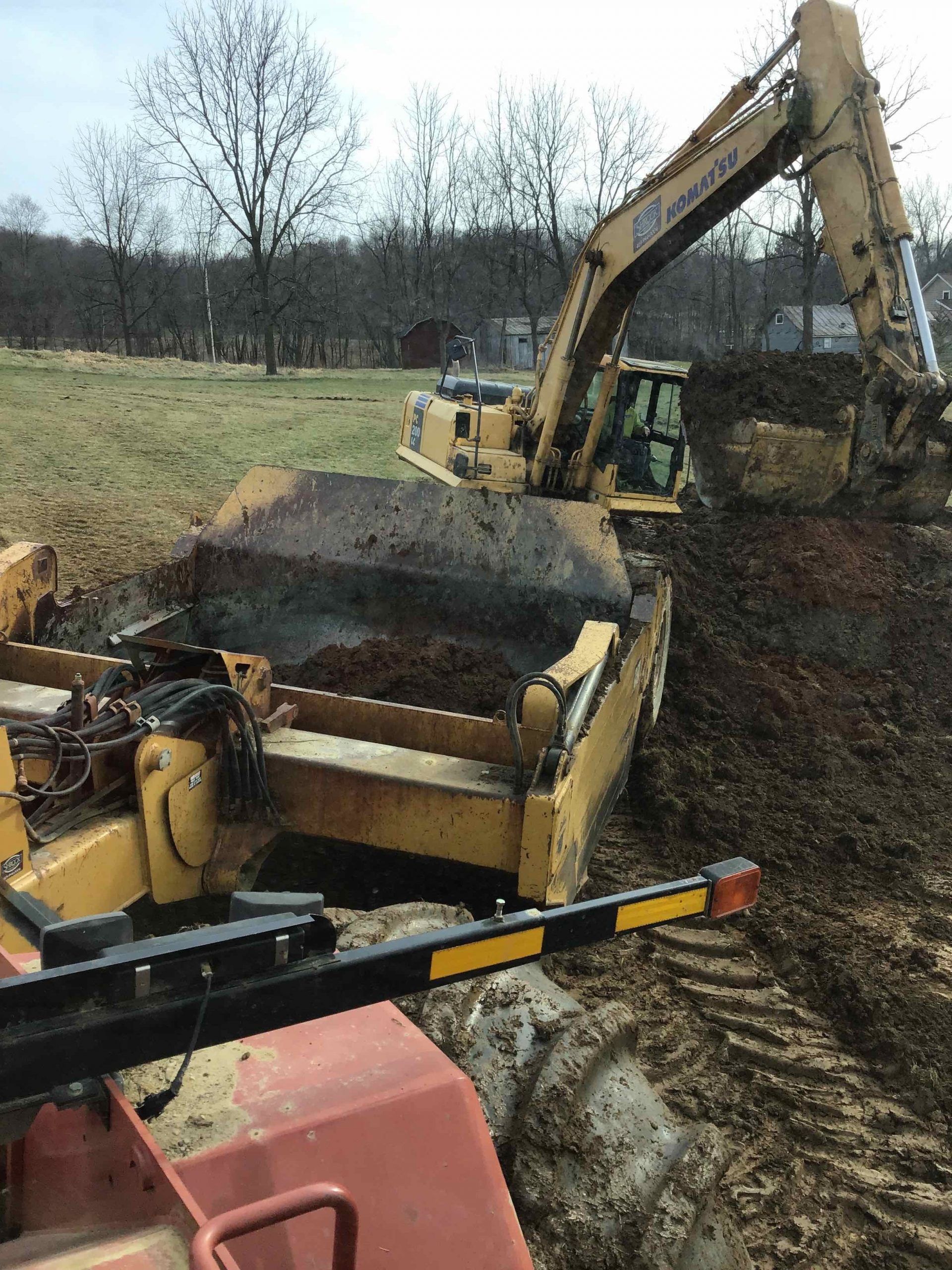 Yellow excavator loading dirt onto a tractor-pulled spreader in a muddy field.