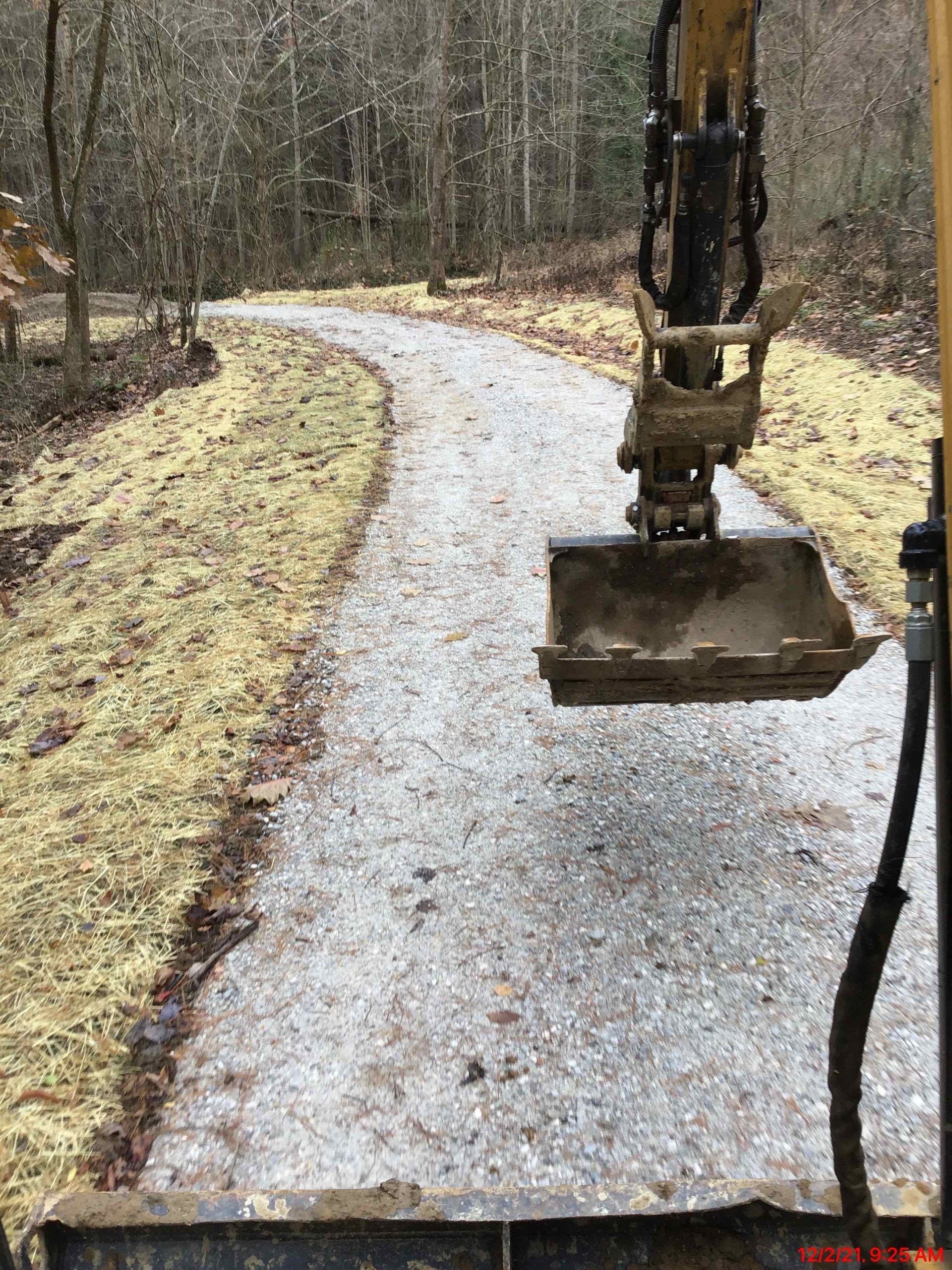 An excavator bucket in view, constructing a gravel path through a wooded area.
