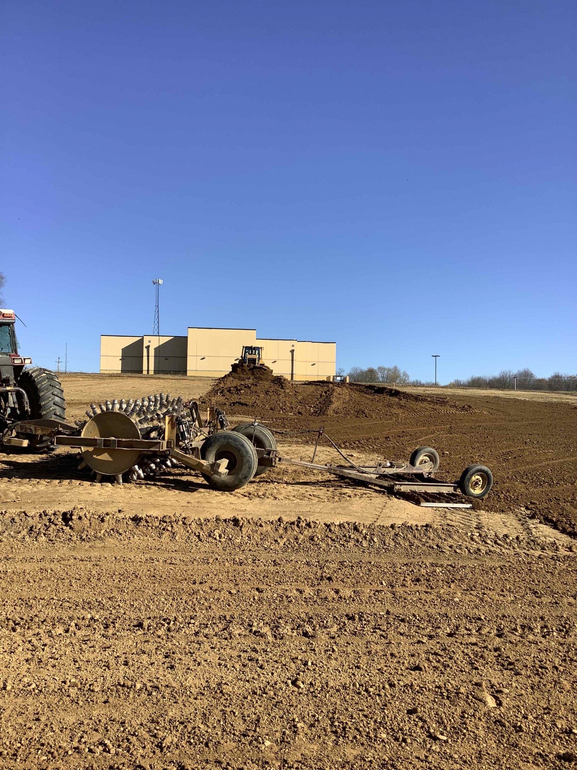 Tractor plowing soil in a field with a small building in the background under a blue sky.
