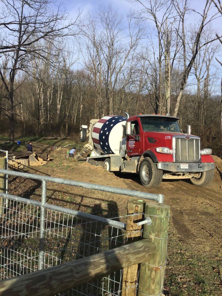 Red cement truck with an American flag design on the barrel, parked on a dirt road near trees.
