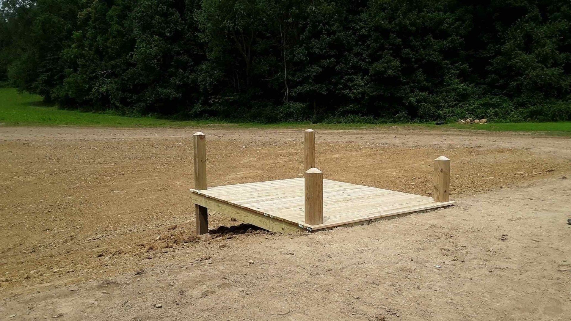 Wooden platform with posts in a gravel area, surrounded by trees.
