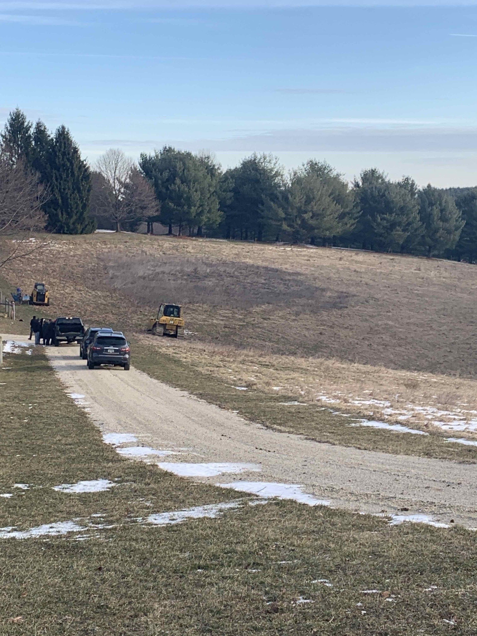 Gravel road through a field; vehicles and construction equipment visible. Trees and blue sky in the background.