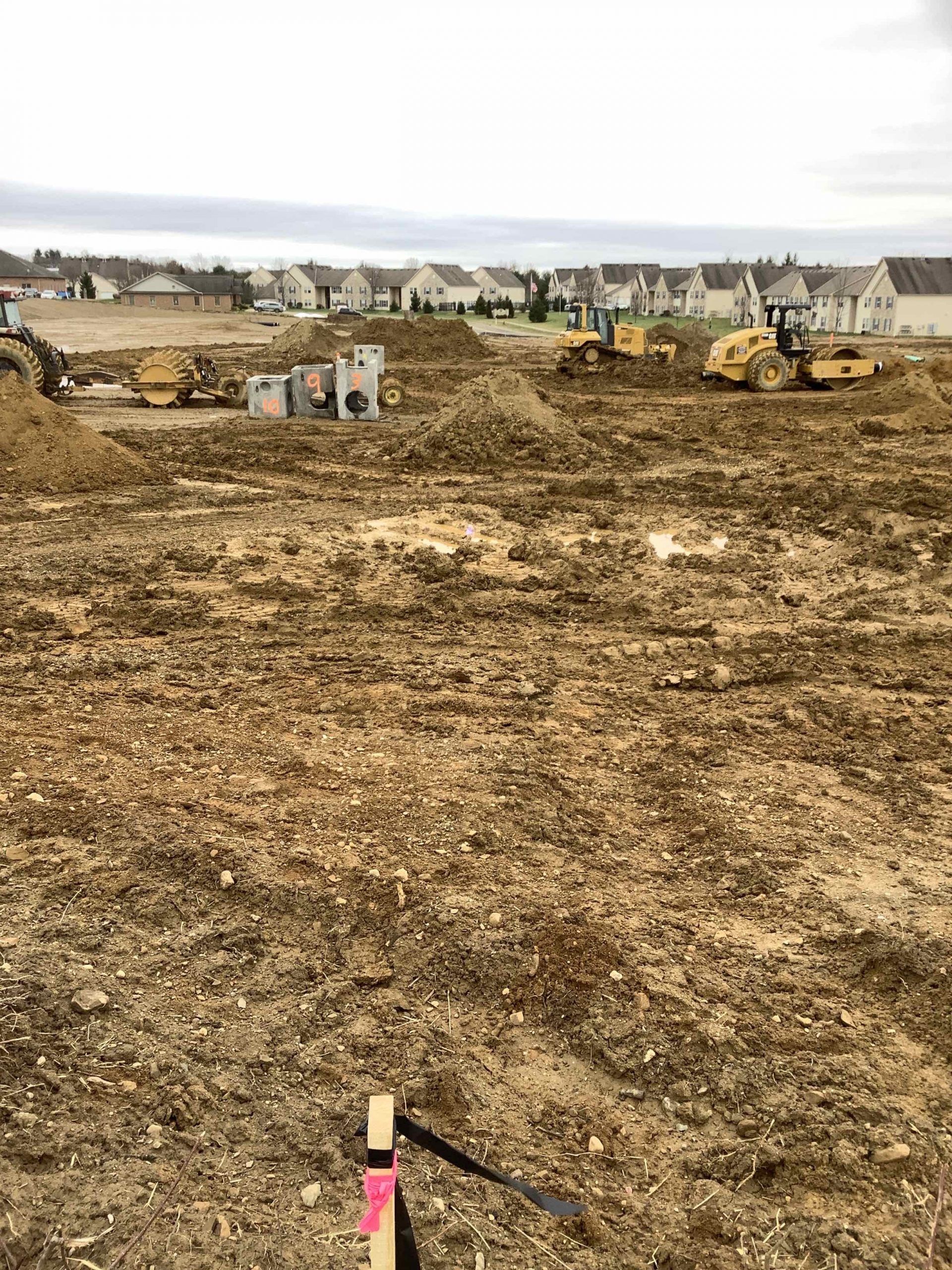 Construction site with heavy machinery, dirt, and houses in the background under a cloudy sky.