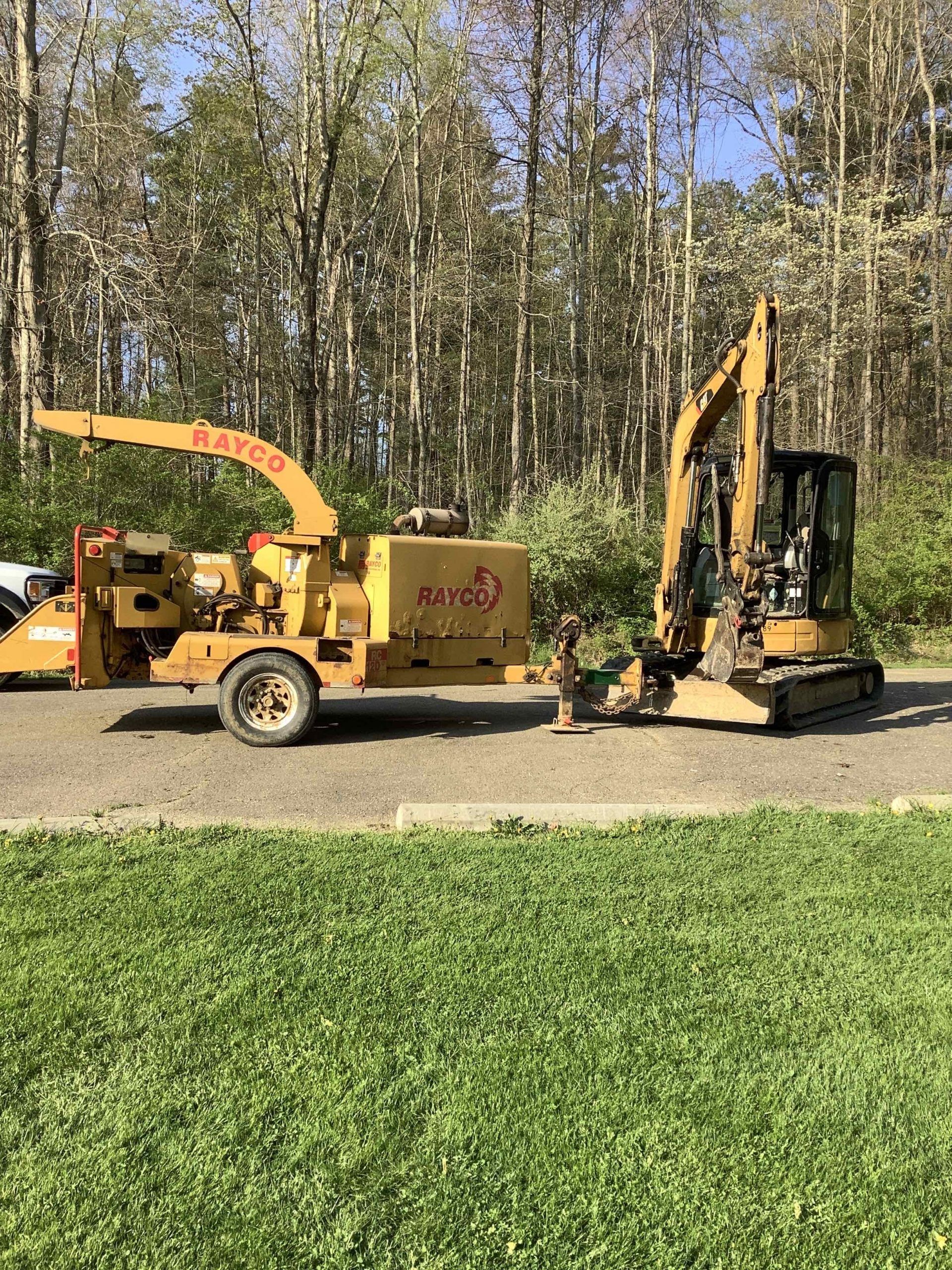 A yellow wood chipper and a small excavator are parked on a paved area, trees in the background.