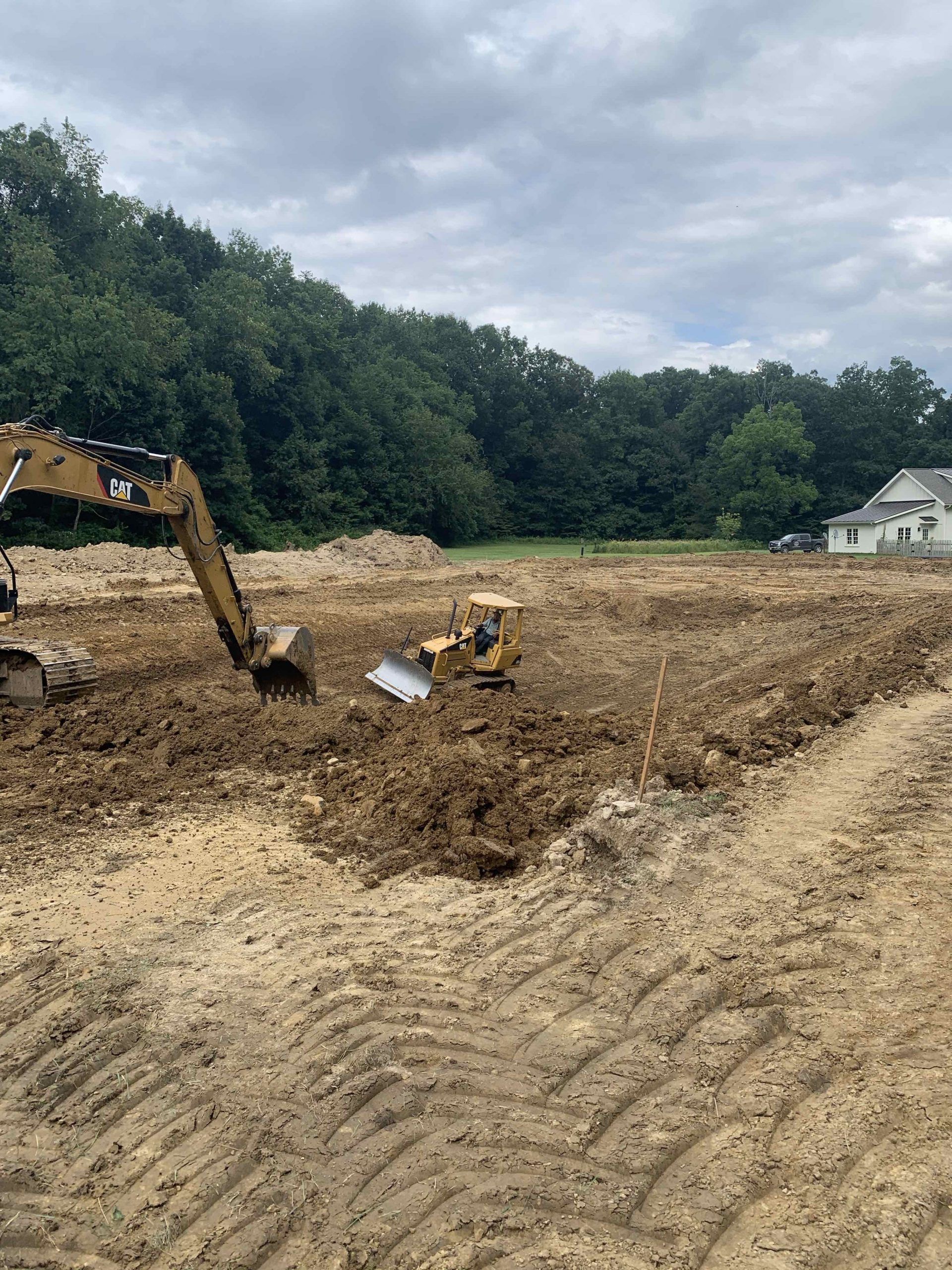 Construction site with excavator and bulldozer digging earth, a house in background, trees and sky.