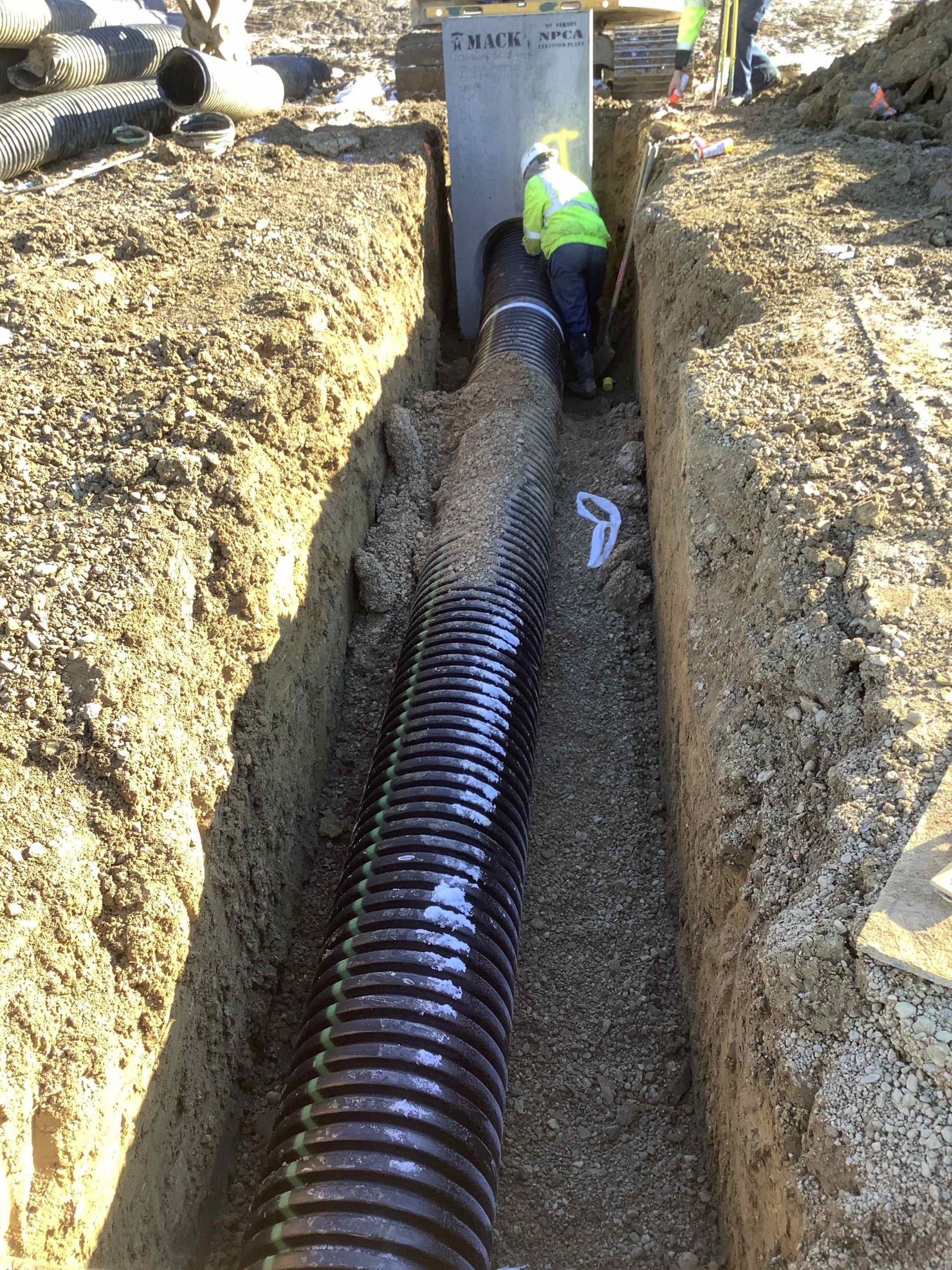 Worker in yellow vest installing black corrugated drainage pipe in a trench.