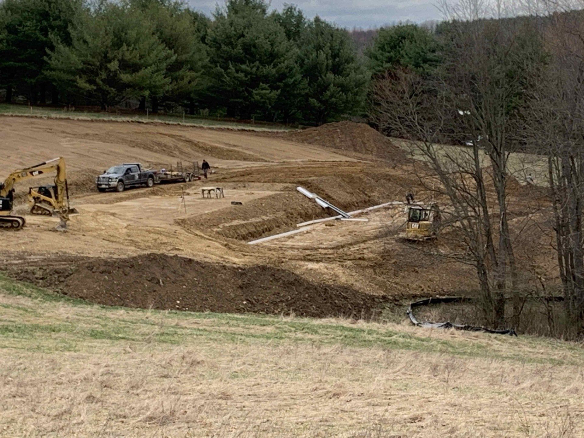 Construction site: Excavators, trucks, and equipment on a dirt field, with trees in the background.